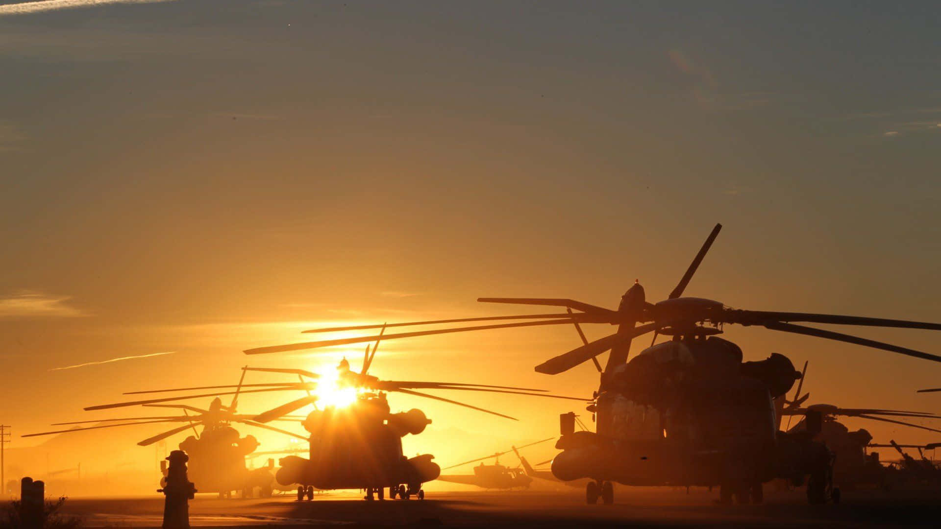 直升機 桌布 壁紙 — 軍用直升機的剪影靜止在停機坪上，夕陽在它們身後落下，散發出金色的光芒。
