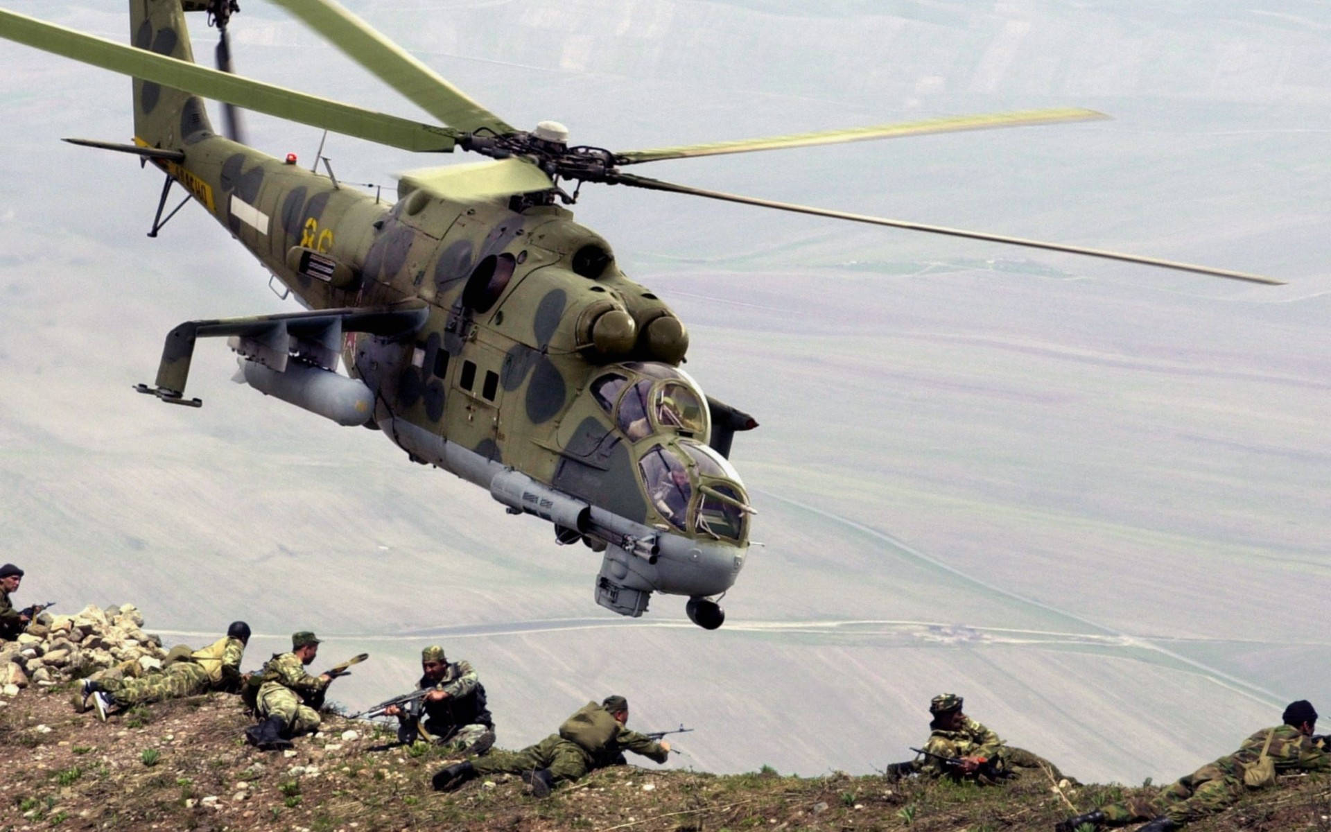 軍用直升機飛越士兵 壁紙 — 一架軍用直升機低空盤旋在身穿迷彩裝備的士兵上方，這些士兵位於岩石山坡上，俯瞰廣闊的山谷。