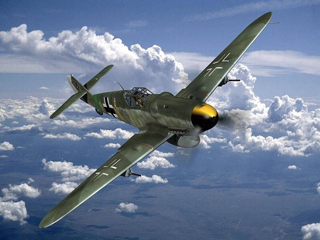 軍綠色Bf 109 德國二戰戰鬥機 壁紙 — 一架綠色二戰戰鬥機，黃色機頭，在明亮的藍天背景下穿過蓬鬆的白雲俯衝。