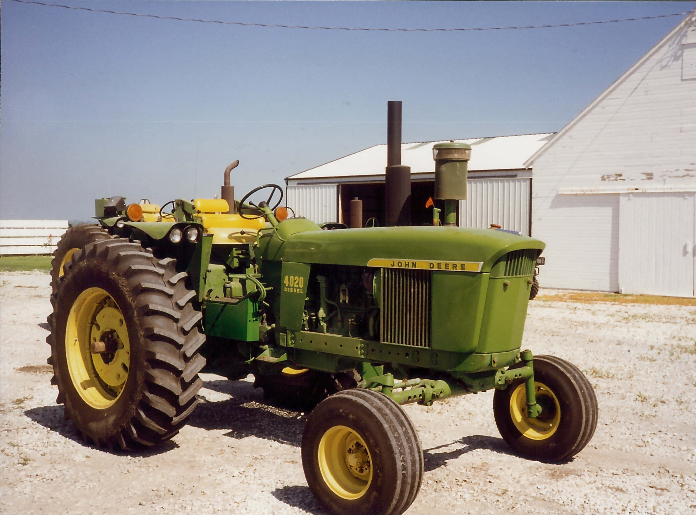 軍綠色拖拉機圖片 壁紙 — 一架復古綠色和黃色的John Deere 4020柴油拖拉機停泊在白色穀倉前的碎石地上，背景是晴朗的藍天。