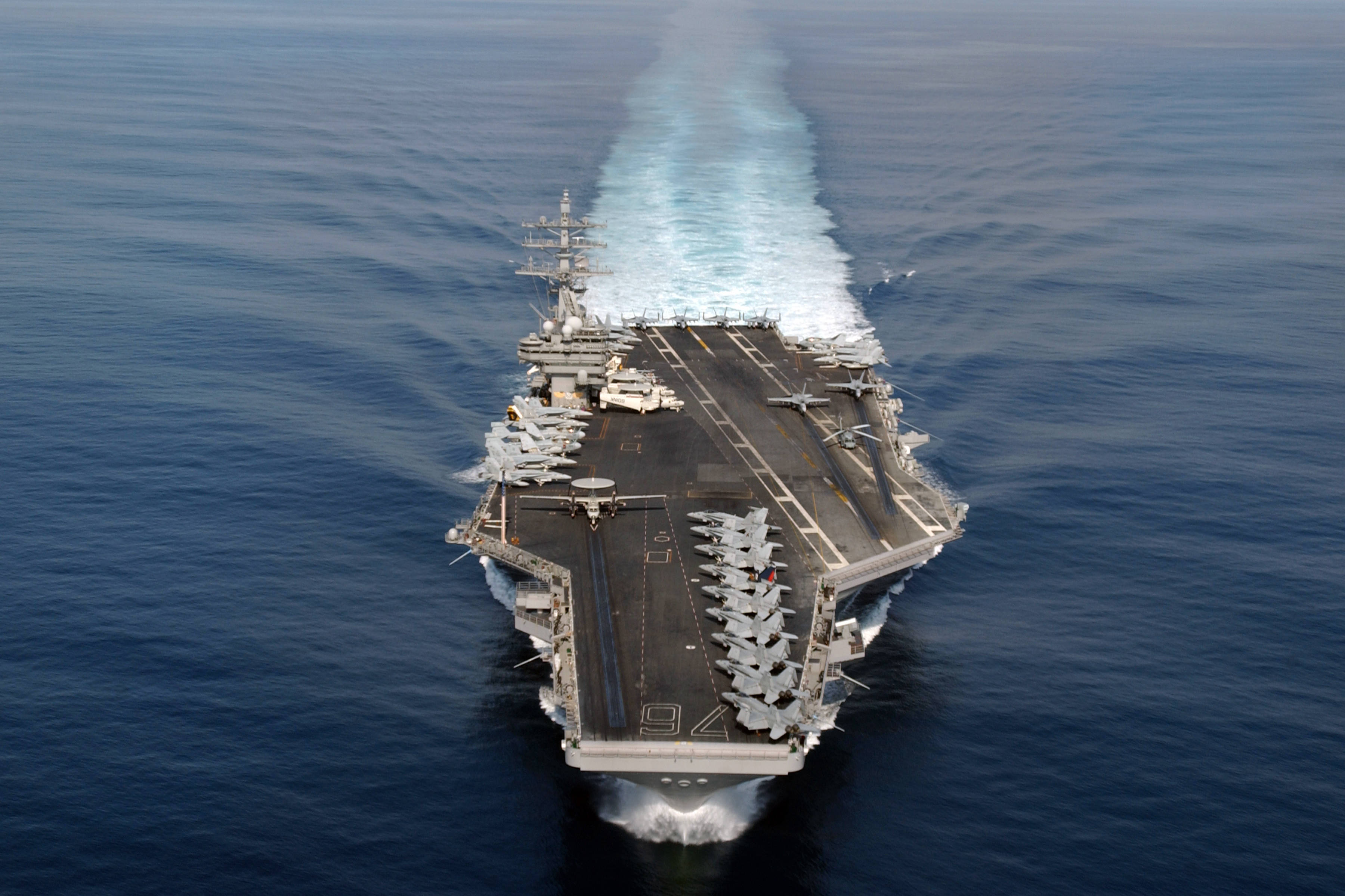 載有戰鬥機的戰艦 壁紙 — 航空母艦劃過深藍色海洋，在其巨大的飛行甲板後留下泡沫般的白色尾跡。