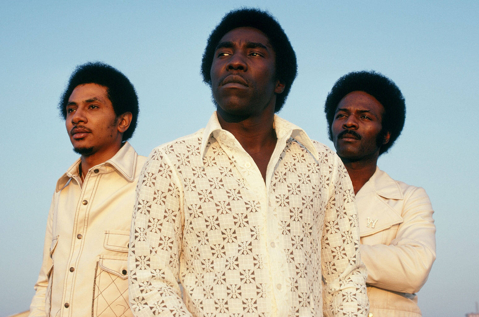 輝煌傳承：Round Hill 收購 O'Jays 音樂作品集 壁紙 — 三位留著非洲爆炸頭、身穿奶油色圖案服裝的男子，在清澈的藍天下向上凝視。