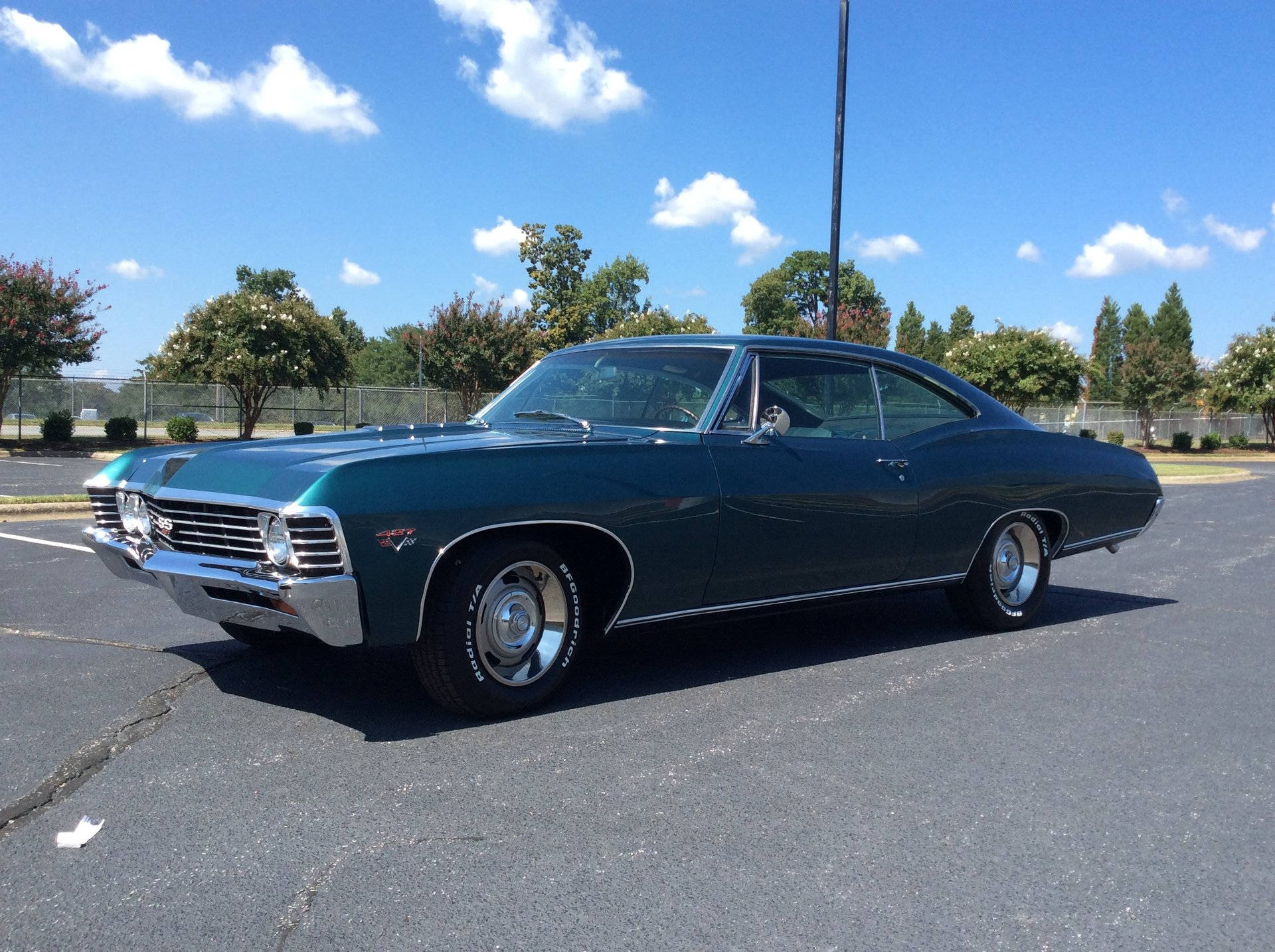 輝煌的寶石綠雪佛蘭Impala 1967 壁紙 — 一輛時尚的深綠色經典肌肉車在晴朗藍天下陽光普照的停車場中閃閃發光。