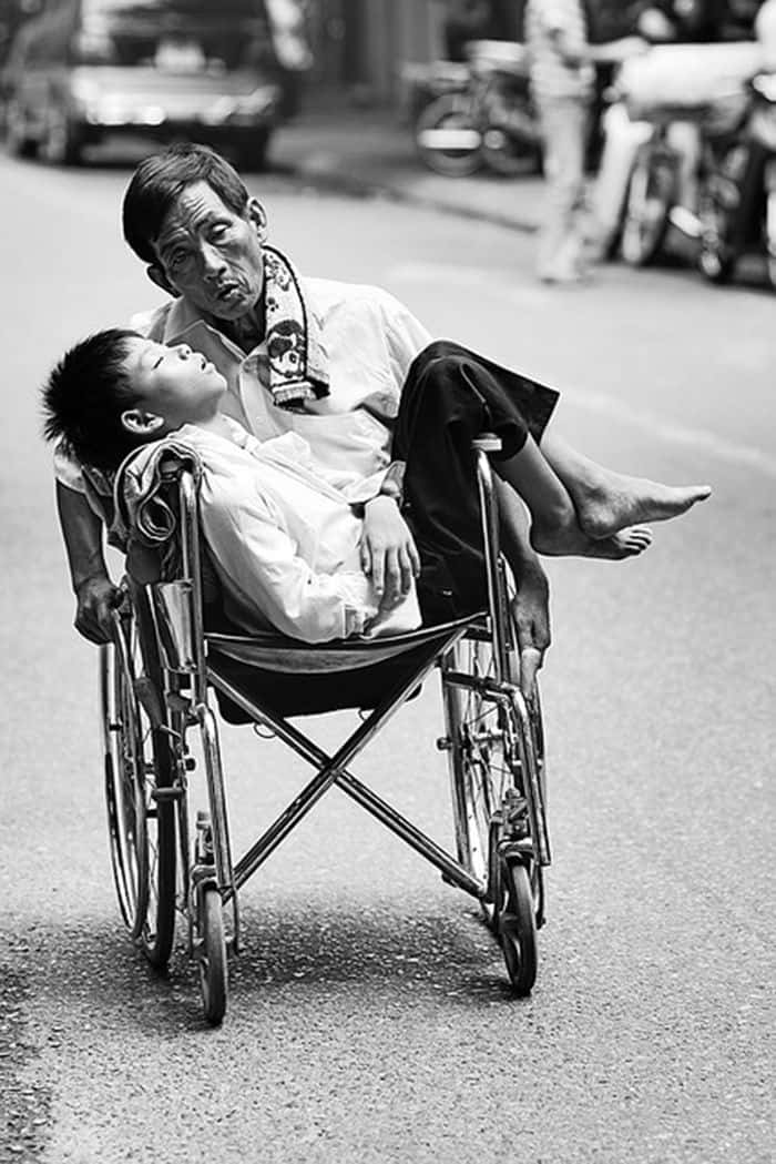 輪椅上的父子情深黑白照片 壁紙 — 一名男子背著孩子，同時推著輪椅沿街前行，黑白照片。