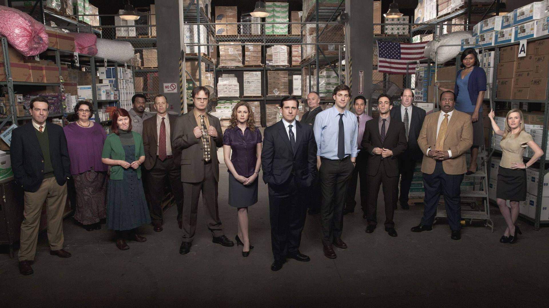 《The Office》演員站在一個有貨架盒子和美國國旗的倉庫中。