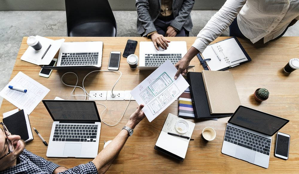 辦公桌上的企業級商業計劃 壁紙 — Overhead view of four professionals collaborating around a wooden table with laptops notebooks and coffee cups.
