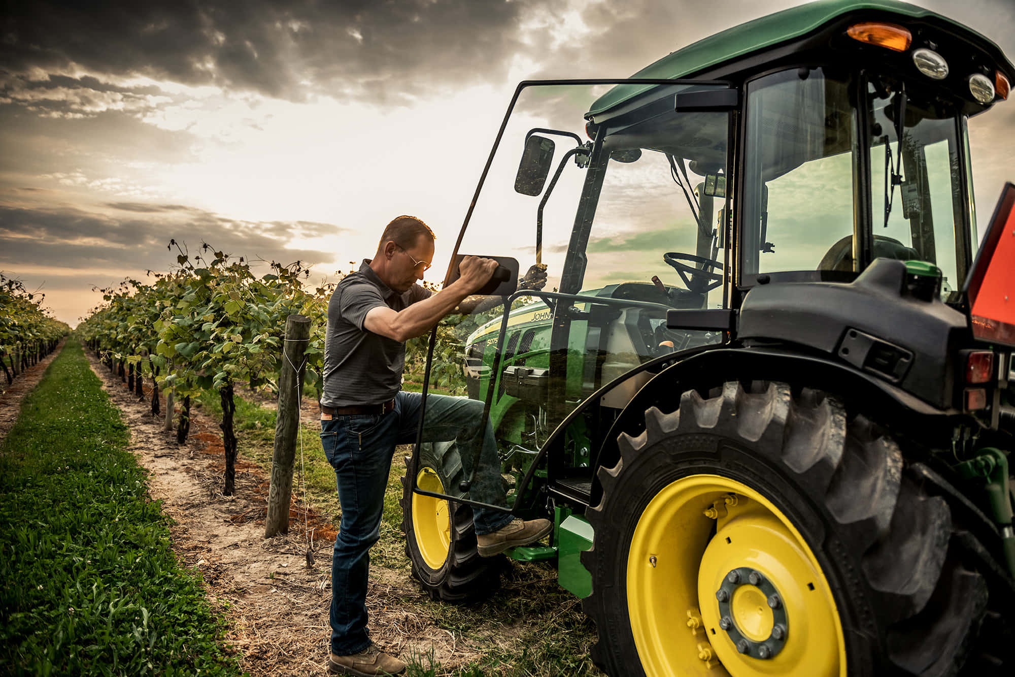 John Deere 桌布 壁紙 — 農夫在日落時分的葡萄園中，在戲劇性的雲層下調整一輛黃色輪子的綠色拖拉機。