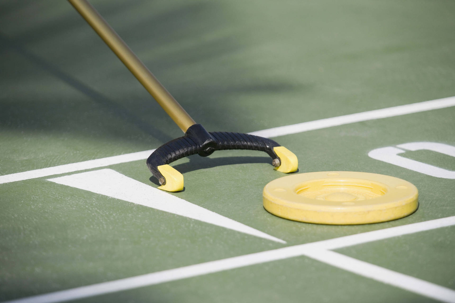 近距離拍攝 Shuffleboard 球與推杆 壁紙 — 一個黃色冰壺石及其手柄的特寫，放置在帶有清晰白色線條的綠色場地上。