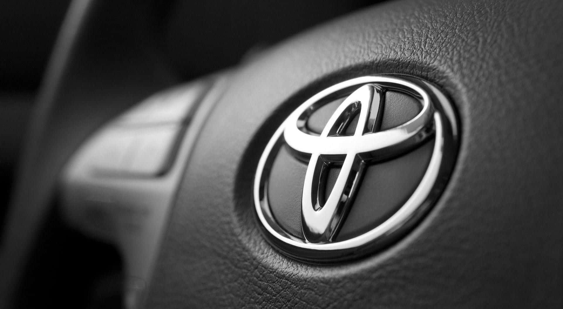 Toyota 桌布 壁紙 — 近距離拍攝一個閃亮的豐田標誌，位於有紋理的黑色皮革方向盤中央，帶有模糊的控制按鈕。