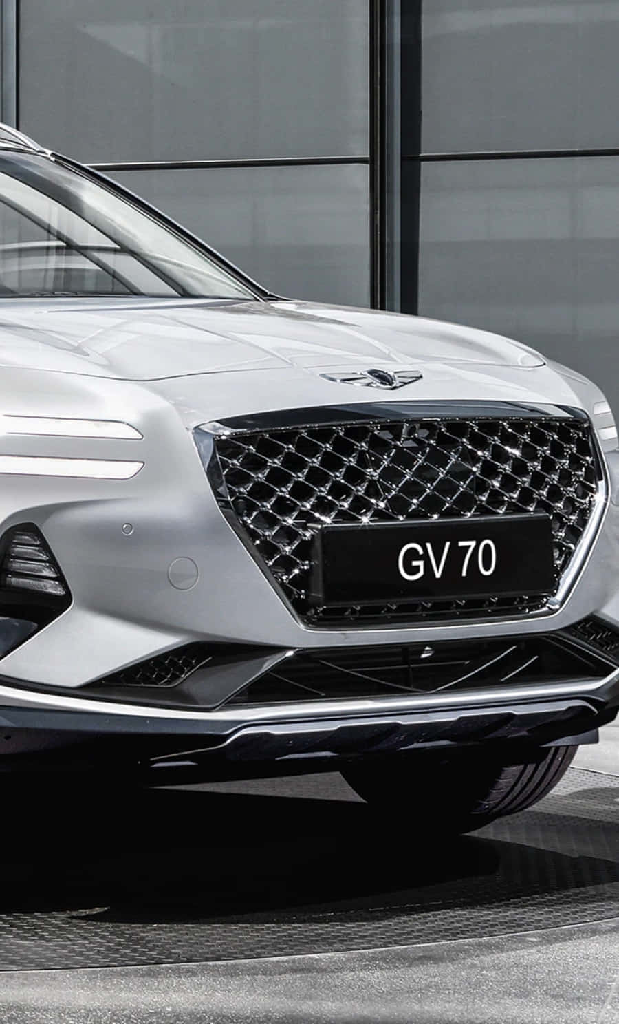 Genesis Gv70 桌布 壁紙 — 近距離拍攝時尚銀色 Genesis GV70 SUV，在玻璃建築背景下展示其大膽的水箱護罩和現代化頭燈。
