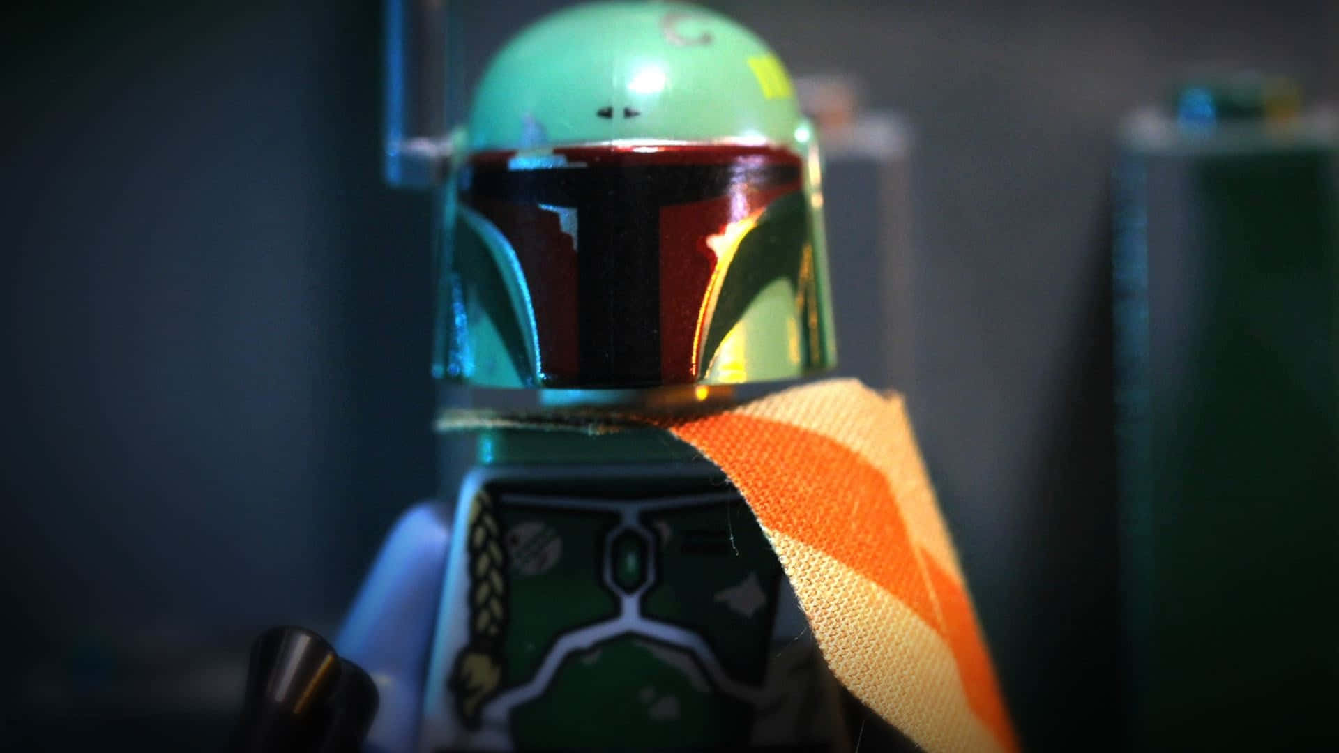Jango Fett 桌布 壁紙 — 近距離拍攝的LEGO波巴·費特人仔，配備反光頭盔和橙色圍巾，背景為暗色模糊效果。