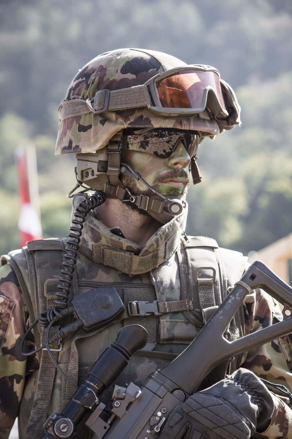 近距離拍攝的軍人制服 壁紙 — 一名身穿全套迷彩裝備和護目鏡的士兵，在模糊的森林背景下持步槍。