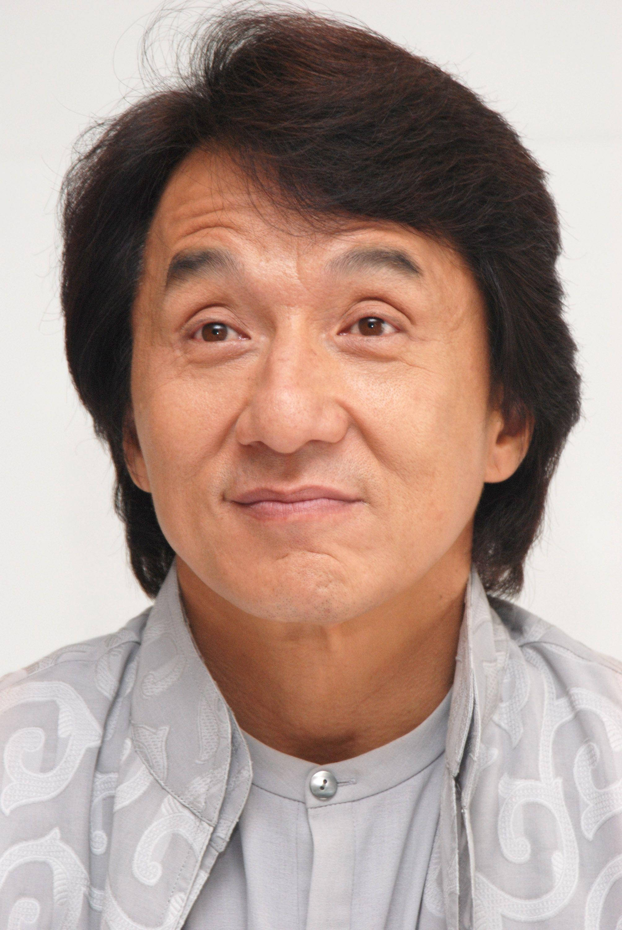 近距離拍攝的驚訝成龍 壁紙 — 一個特寫肖像，一名深色頭髮、帶著溫柔微笑的男子，穿著灰色圖案襯衫，背景為純白色。