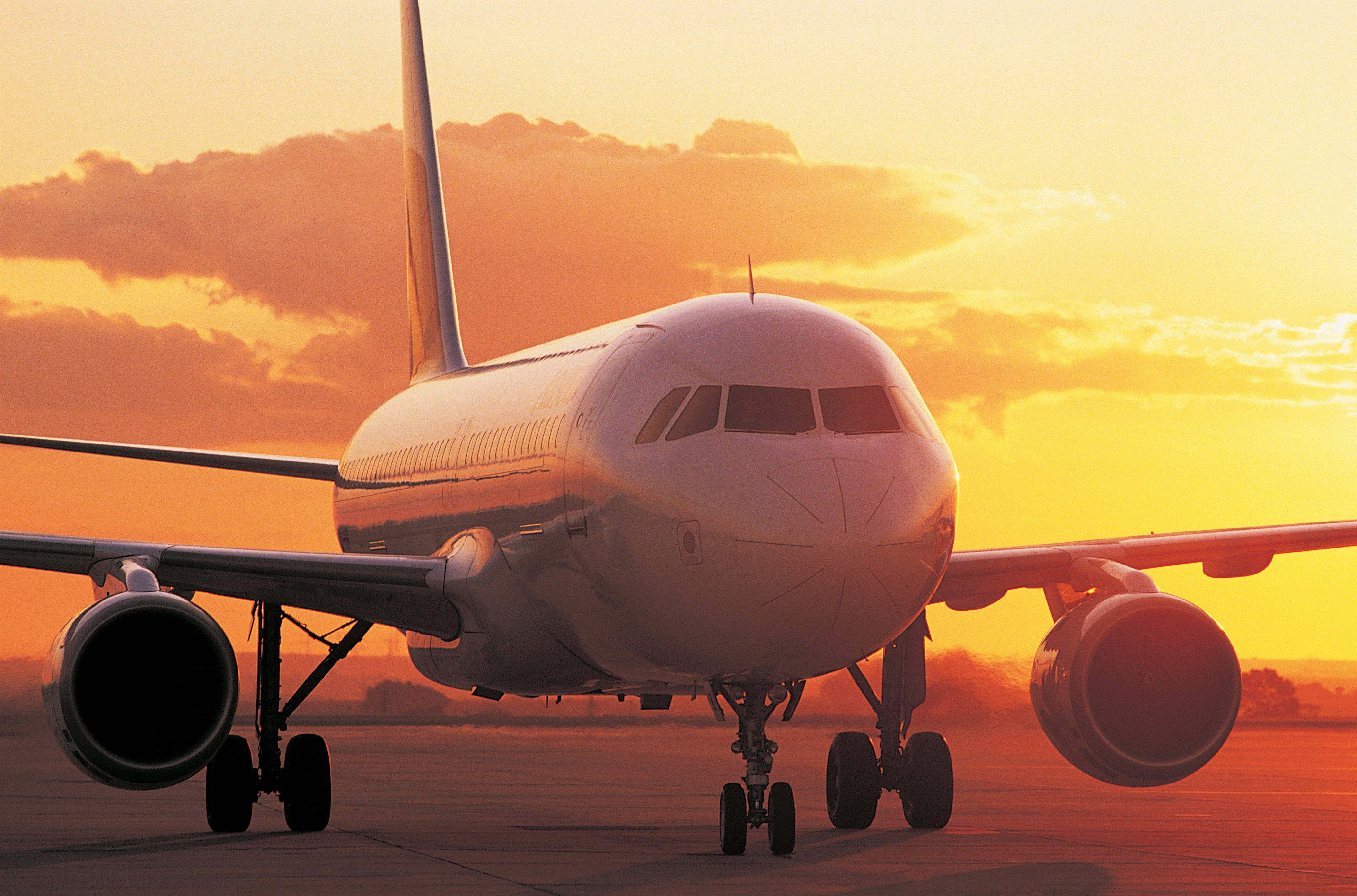 近距離日落飛機背景 壁紙 — 一架商用飛機停在跑道上，沐浴在溫暖的金色日落光線中，上方有戲劇性的橙色雲彩。
