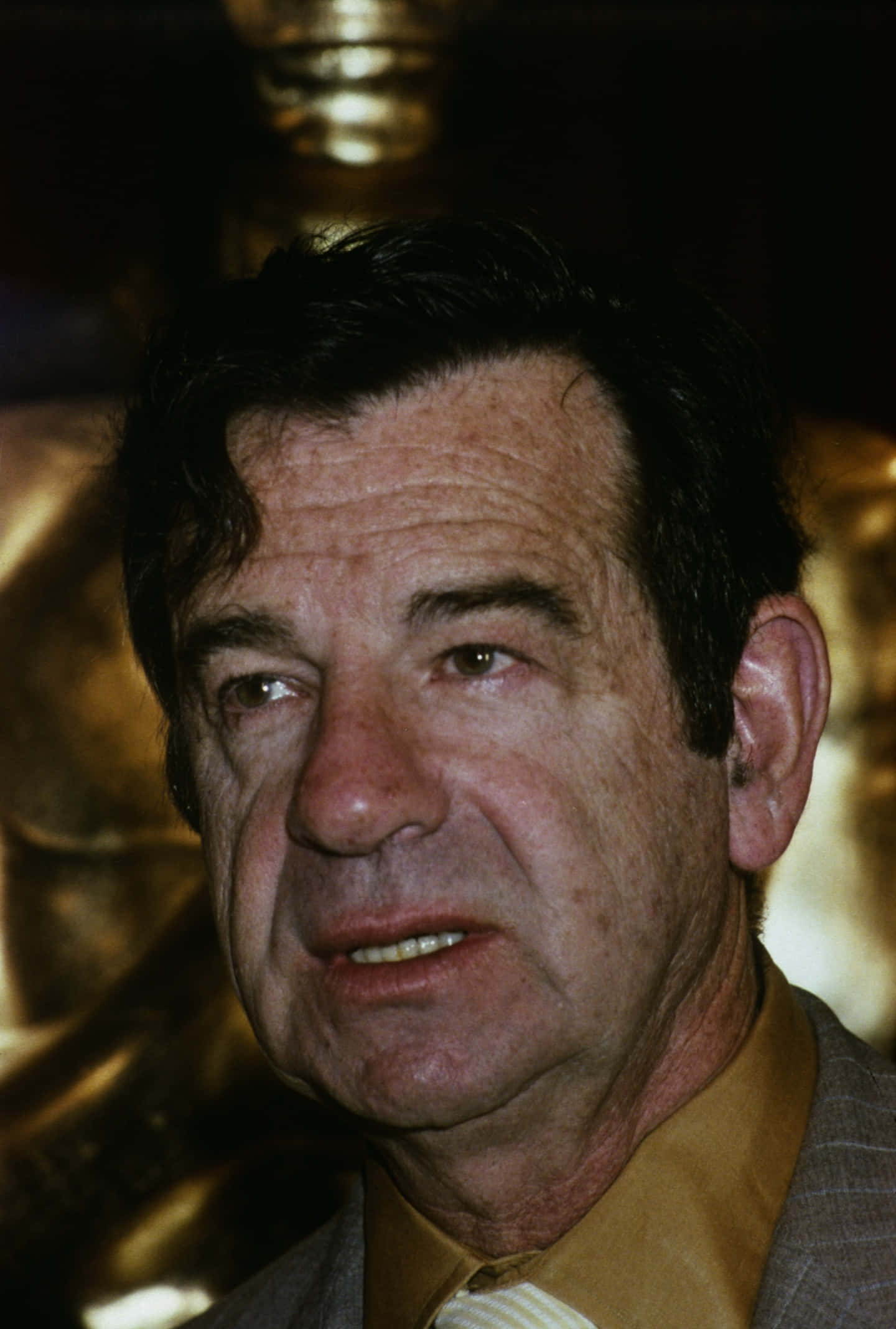Walter Matthau 桌布 壁紙 — 近距離肖像，一名深色頭髮、有皺紋的男子，穿著黃色襯衫和西裝外套，背景是金色雕像。