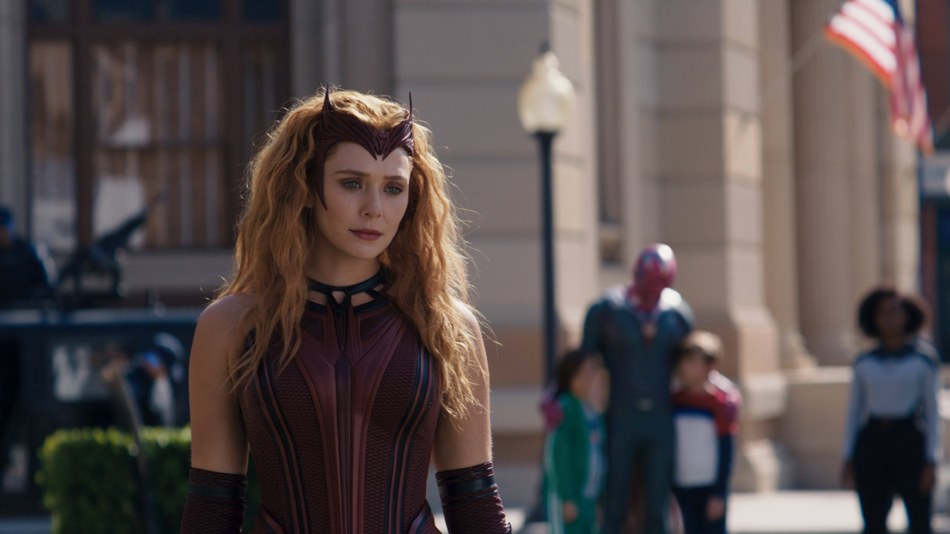 迪士尼+ 旺達終章 壁紙 — Wanda Maximoff 身穿紅黑女巫服裝，在陽光明媚的城市街道上清晰可見，背景有模糊的行人和美國國旗。