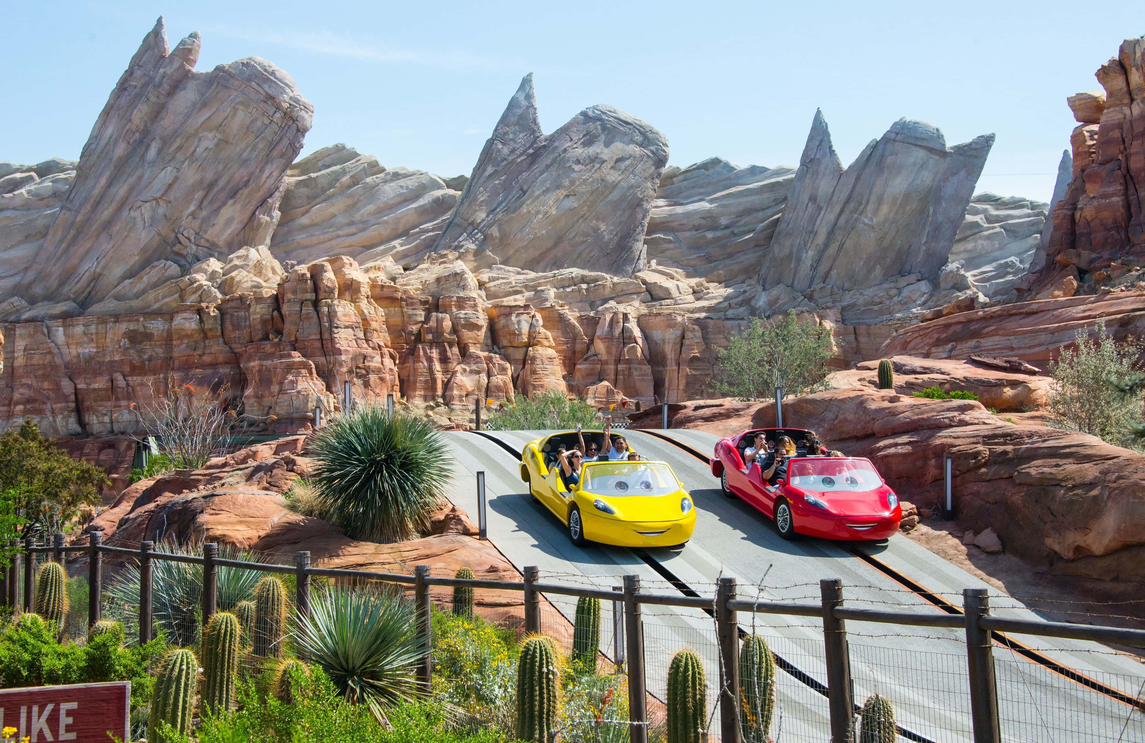 迪士尼樂園 Radiator Spring Racer 主題背景 壁紙 — 兩架色彩繽紛的動畫汽車在晴朗藍天下，沿著被岩石懸崖和仙人掌包圍的沙漠賽道奔馳。