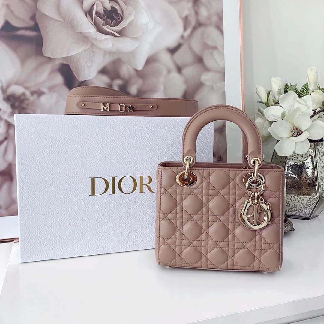 迪奧的經典優雅 壁紙 — 一款粉紅色Dior Lady Dior手袋，配以金色金屬配件，優雅地放在其品牌盒子和白色花朵旁邊，置於潔白表面上。