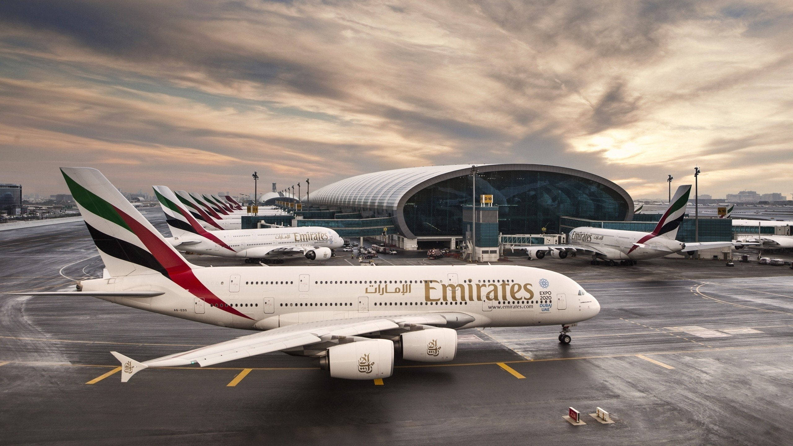 迪拜國際機場的空中巴士 壁紙 — 阿聯酋航空A380客機在杜拜機場航站樓前排成一排，背景是戲劇性的金色日落天空。