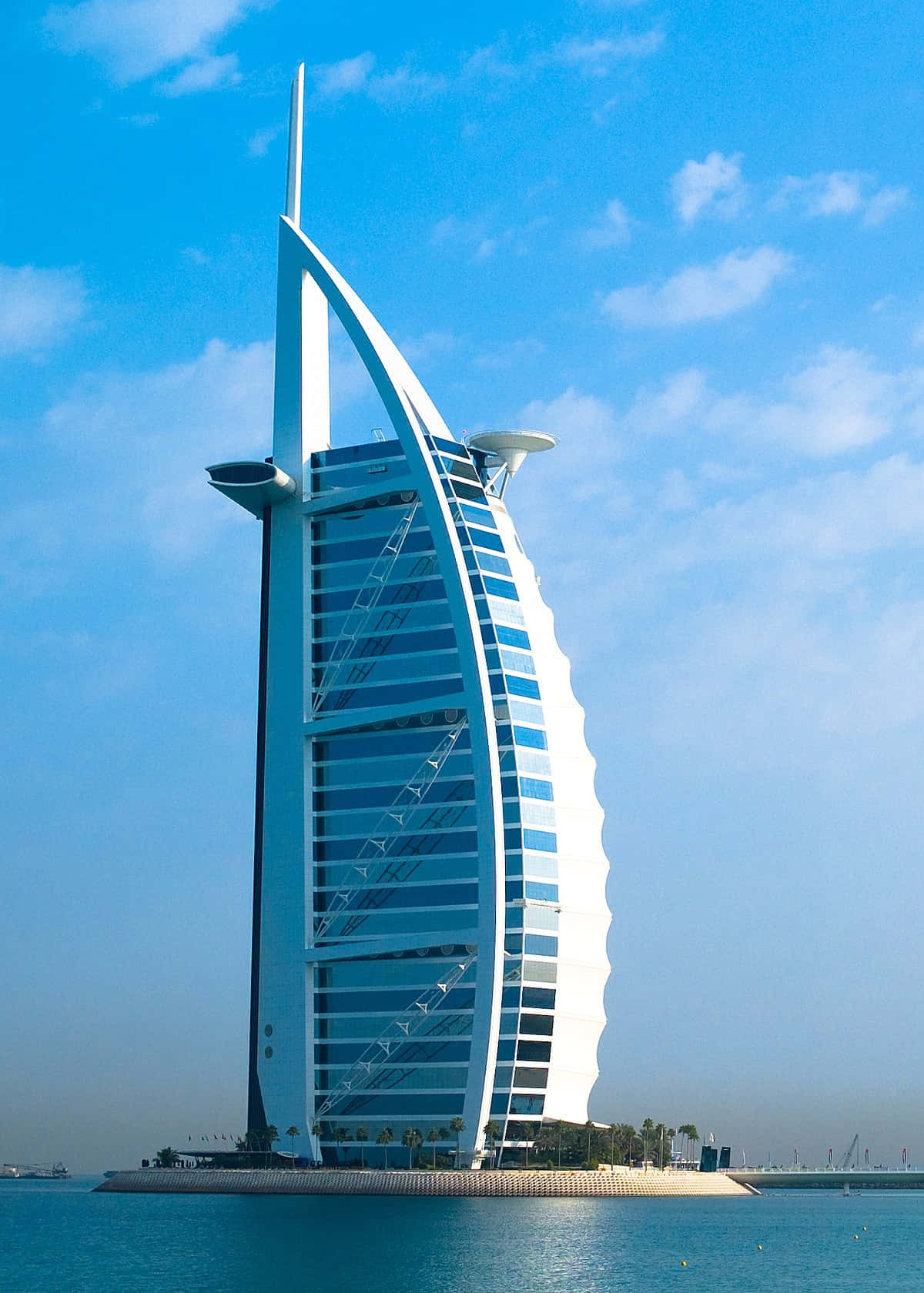 迪拜夜幕下超現代帆船酒店 壁紙 — 帆船形狀的 Burj Al Arab Jumeirah 酒店在蔚藍天空和零星白雲下閃耀，玻璃外牆將陽光反射在平靜的碧綠水面上。