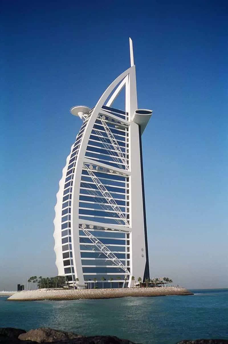 迪拜帆船酒店暮色美景 壁紙 — 帆船形狀的帆船酒店（Burj Al Arab Jumeirah）在清澈的藍天下高聳，白色外牆在碧綠的海水上反射陽光。