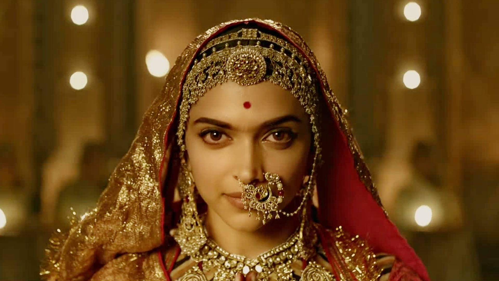 迪皮卡·帕度柯妮《Padmaavat》歌劇舞造型 壁紙 — 一位身穿華麗金色新娘禮服、戴著紅色頭紗和精緻珠寶的女性，在溫暖閃爍的燈光下向前凝視。