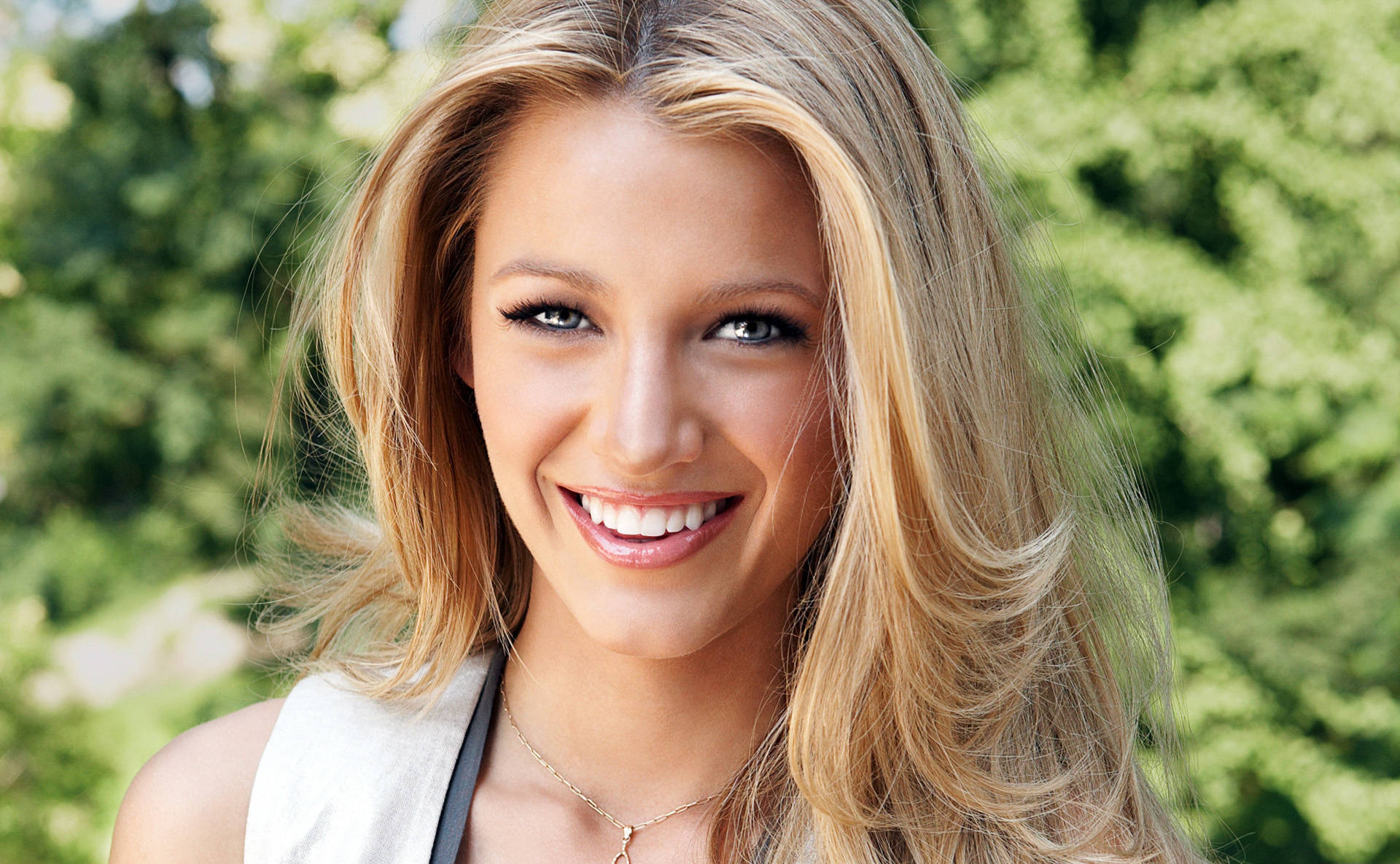 迷人 Blake Lively 壁紙 — 一位容光煥發的金髮女性在茂密的綠色戶外背景下，柔和自然光線中溫暖地微笑。