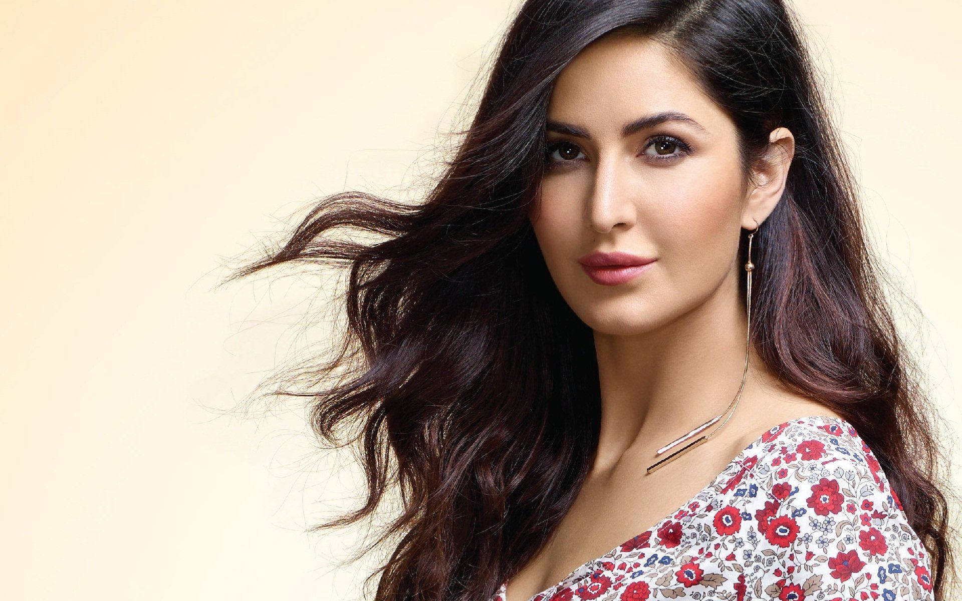 迷人 Katrina Kaif 壁紙 — 一位擁有飄逸深色長髮、身穿花卉圖案上衣的女性，在柔和米色背景前自信地凝視。