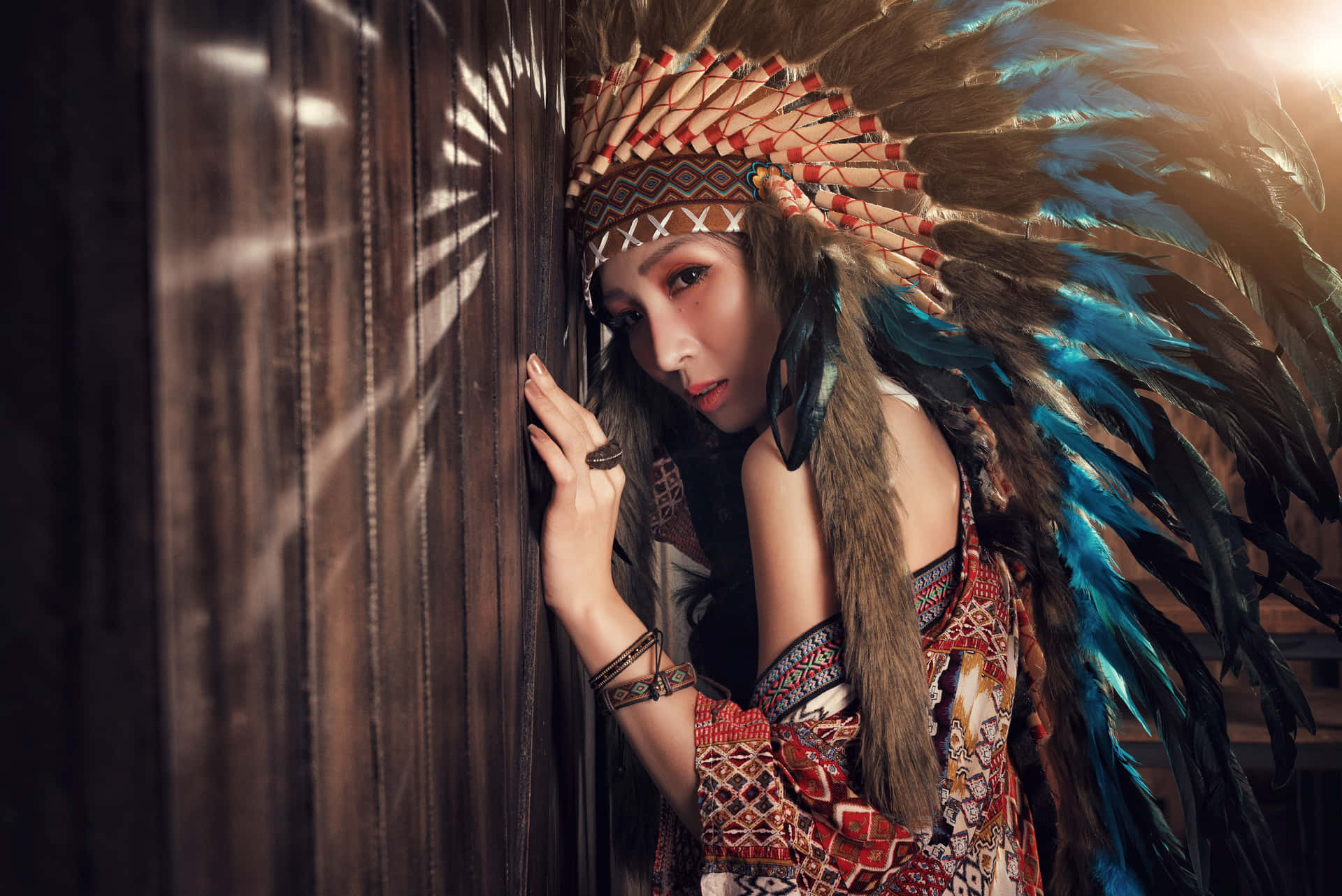 一位戴著傳統羽毛頭飾的女人靠在一扇木門上，陽光照射進來。