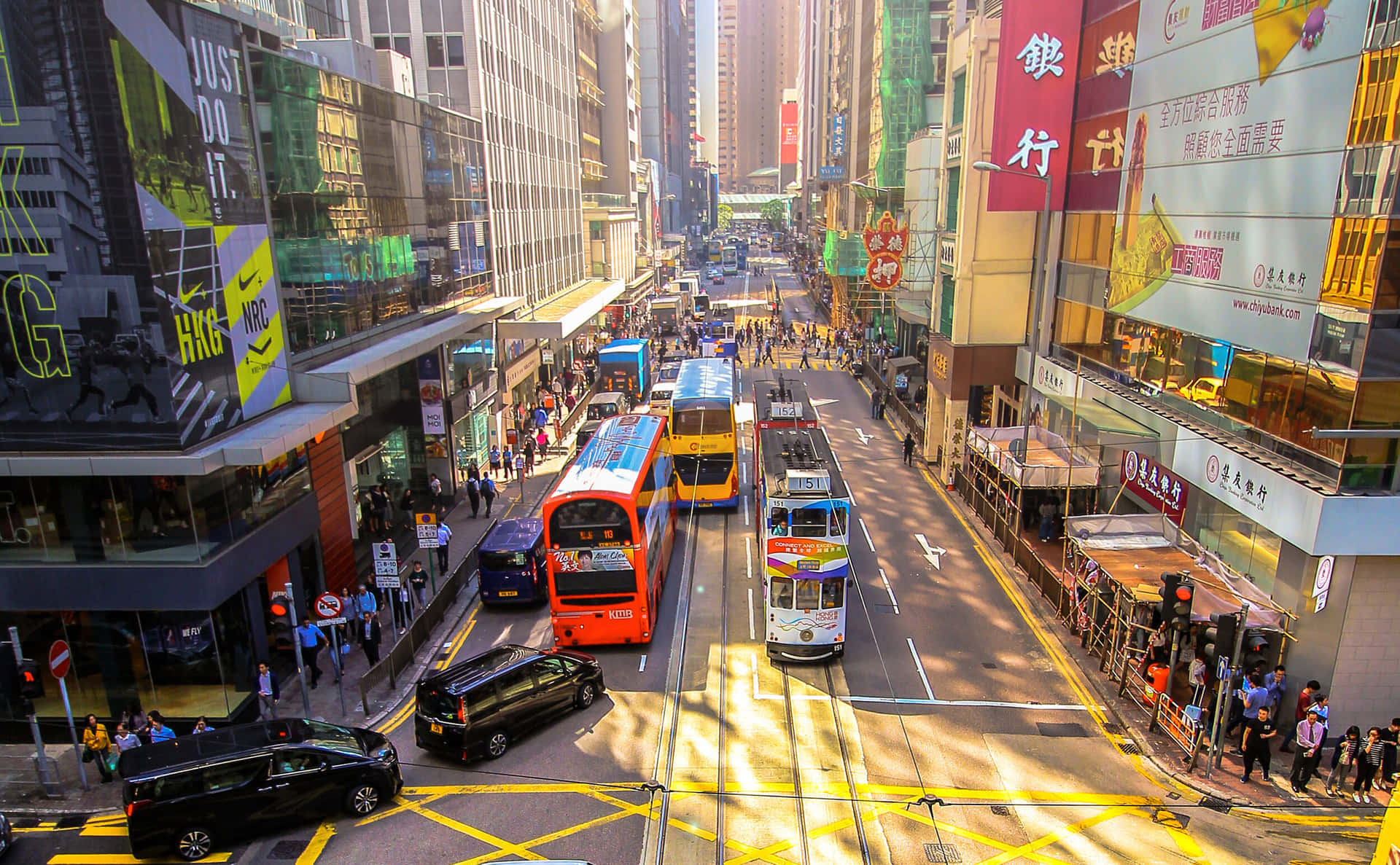迷人夜景中的香港天際線 壁紙 — 一條繁忙的香港街道，充滿色彩繽紛的雙層巴士、電車和行人，在明亮的陽光下投下長長的影子。