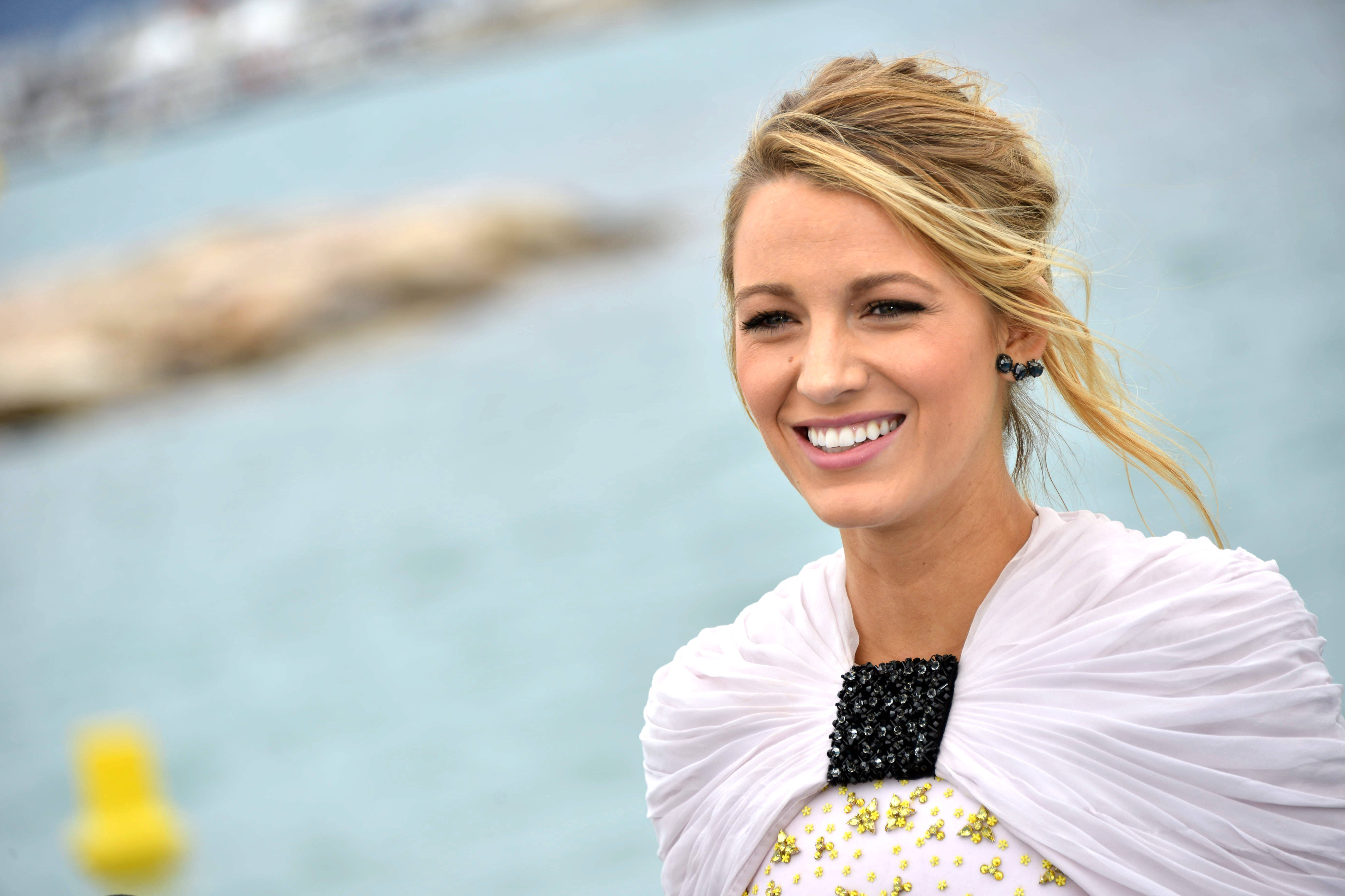 迷人微笑的 Blake Lively 壁紙 — 一位微笑的金髮女性，身穿白色垂墜服飾，帶有黑色珠飾細節，站在模糊的藍色海洋和岩石海岸前。