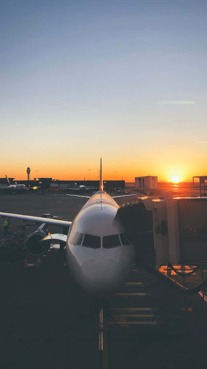 迷人旅程：旅行的優雅風采 壁紙 — 一架客機停靠在機場閘口，夕陽在橙色和藍色的漸變天空中落下。