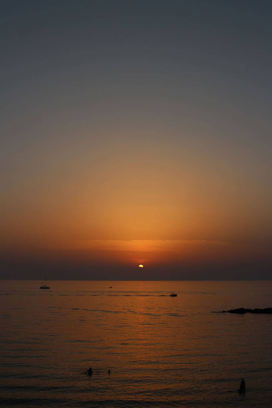 夕陽映照海洋，船隻和游泳者剪影映襯橙色天空