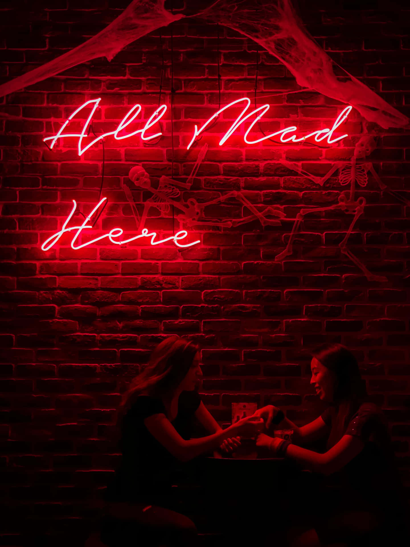 兩位女士在寫著 All Mad Here 的紅色霓虹燈牌下玩牌。