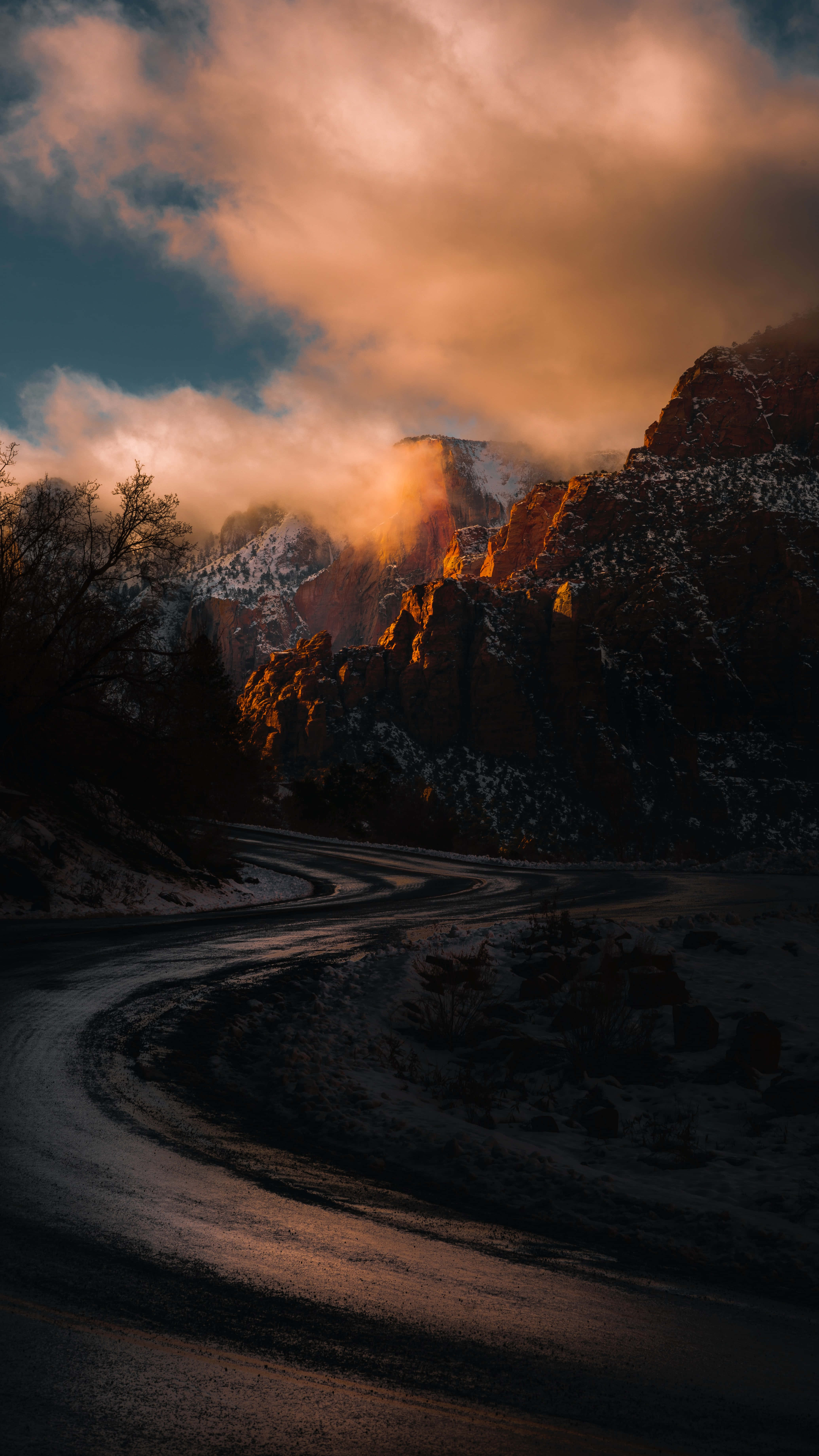 迷人暗色彎道與遠山iPhone背景 壁紙 — 一條蜿蜒的雪路穿過戲劇性的山口，金色夕陽穿透雲層，照亮積雪的山峰。