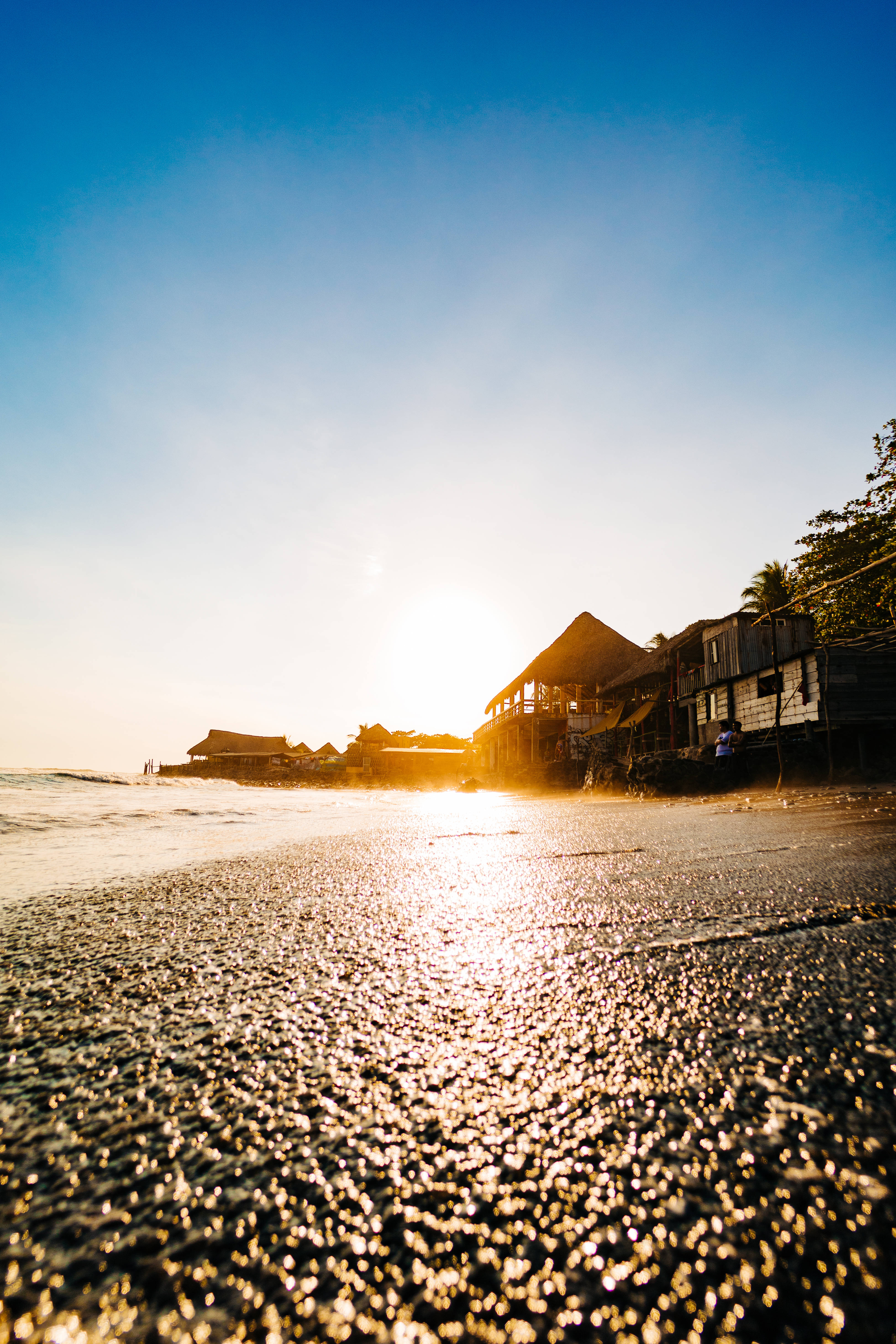 迷人海岸風光：薩爾瓦多海灘天堂 壁紙 — 夕陽在濕沙上閃爍，木製海灘小屋在清澈的藍天下形成剪影。