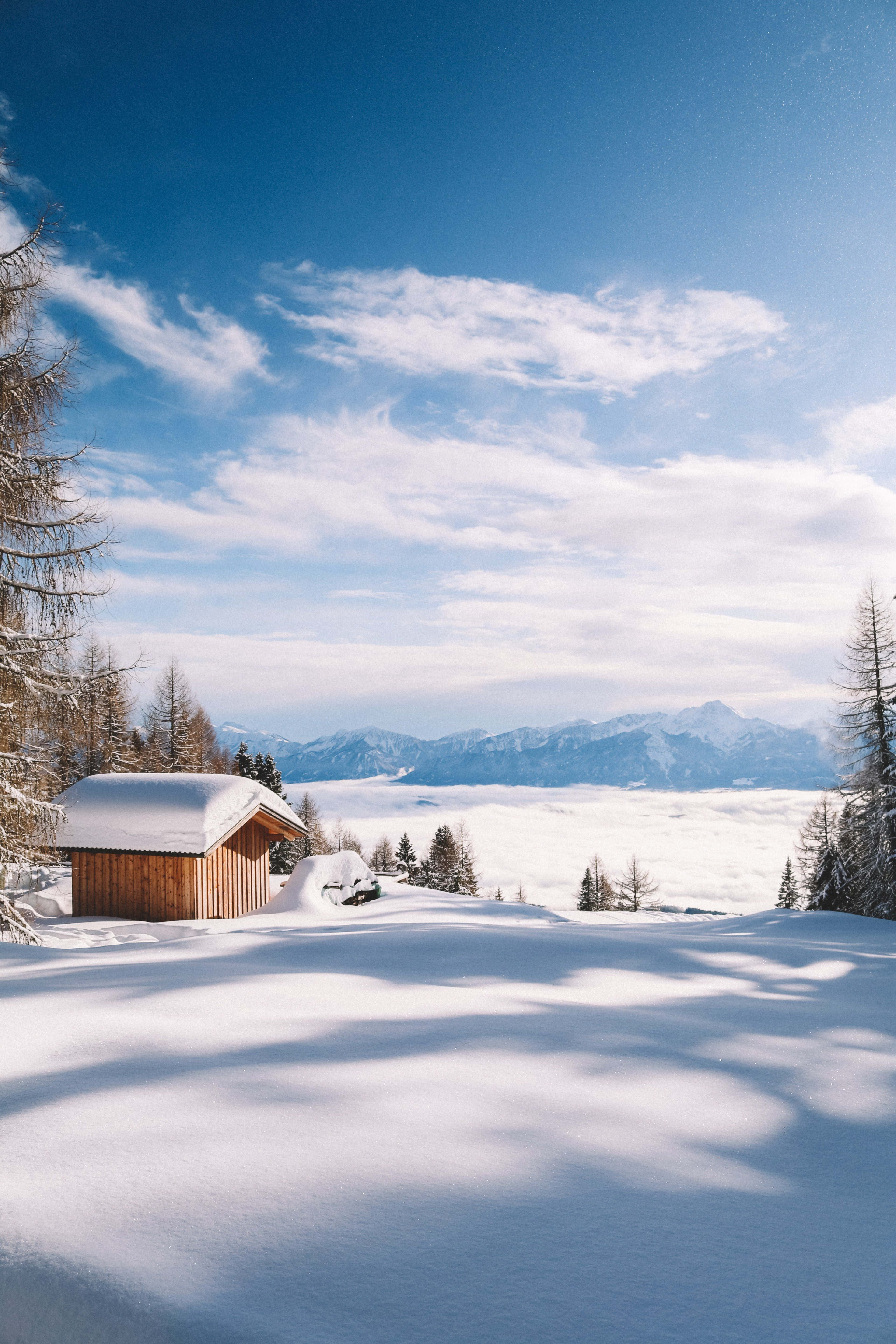 迷人的冬季仙境風景 壁紙 — 一間木屋坐落在積雪覆蓋的山脈中，天空湛藍，雲朵稀疏。