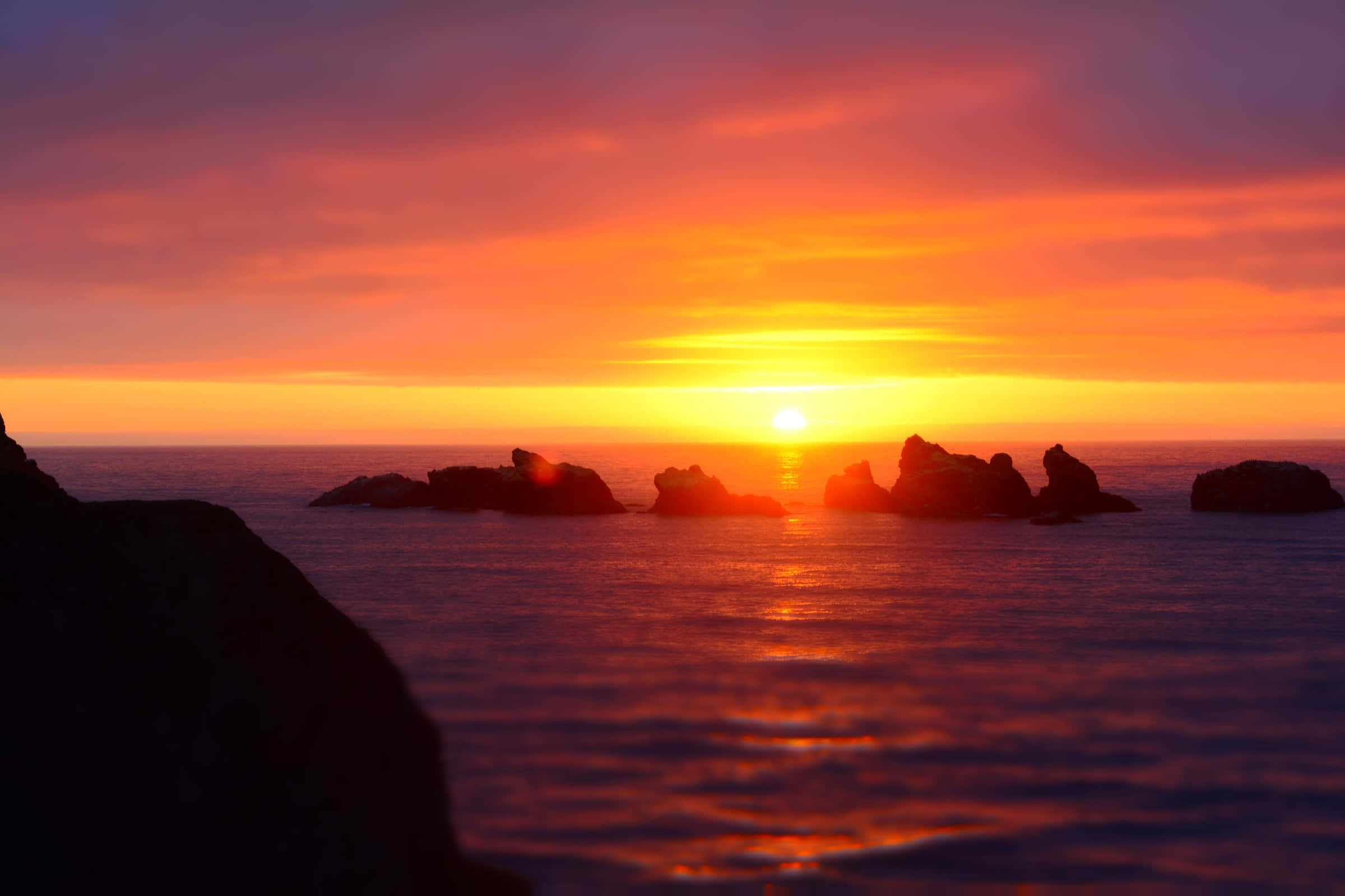 迷人的地平線日落景觀 壁紙 — 熾熱的夕陽將海洋染成橙色和紫色，剪影般的岩石矗立在發光的地平線上。