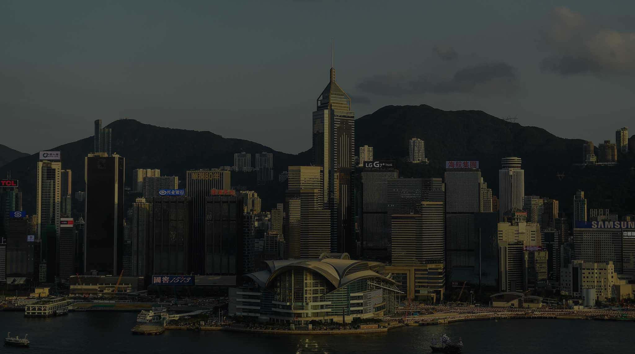 迷人香港天際線夜景 壁紙 — 黃昏時分，香港天際線燈光昏暗，高聳的摩天大樓和山脈作為背景。