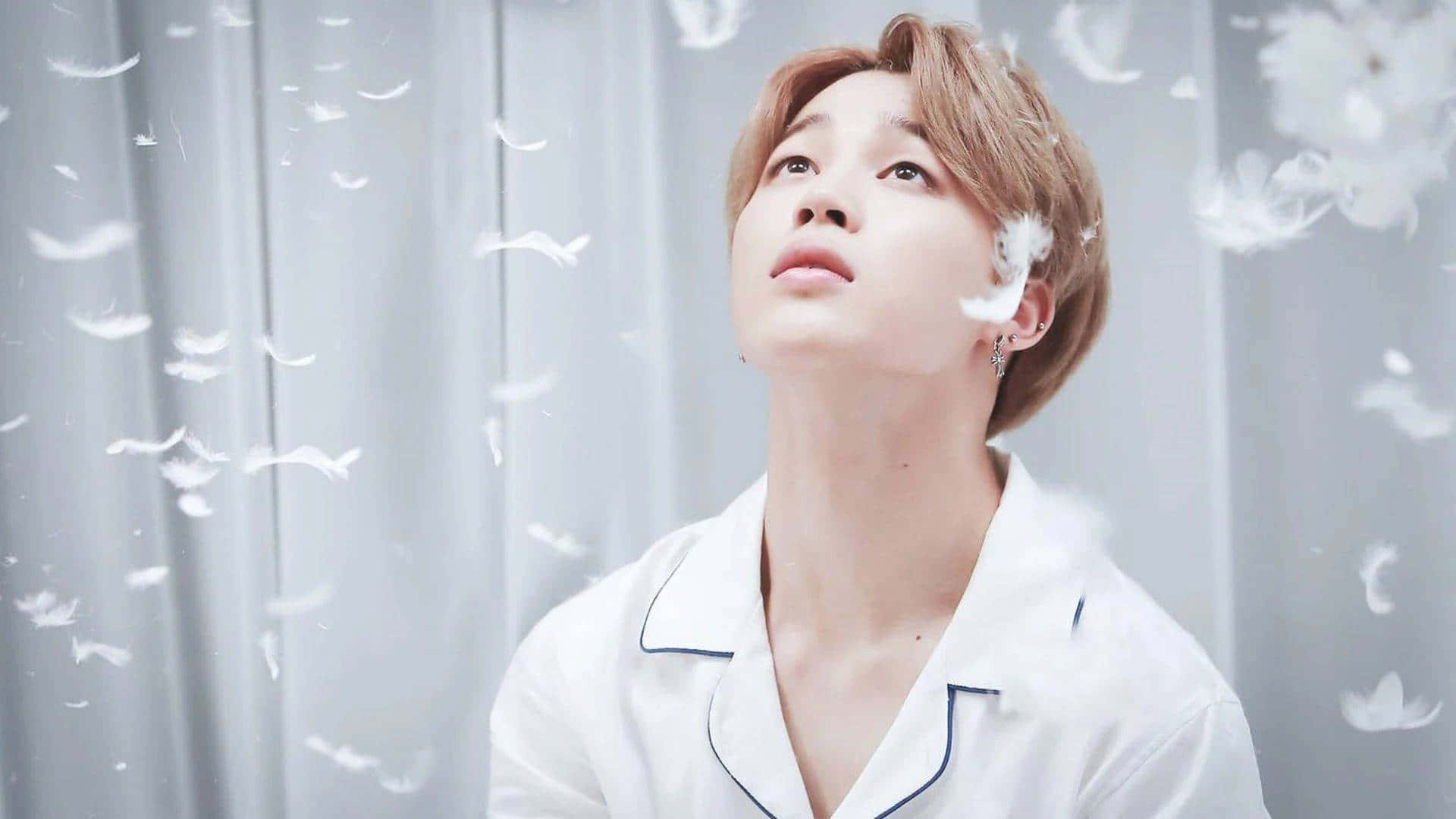 迷人魅力：BTS Jimin 的迷人姿態 壁紙 — 一名身穿白色睡衣的年輕男子向上凝視，柔軟的白色羽毛在他周圍飄動，背景是淺灰色窗簾。