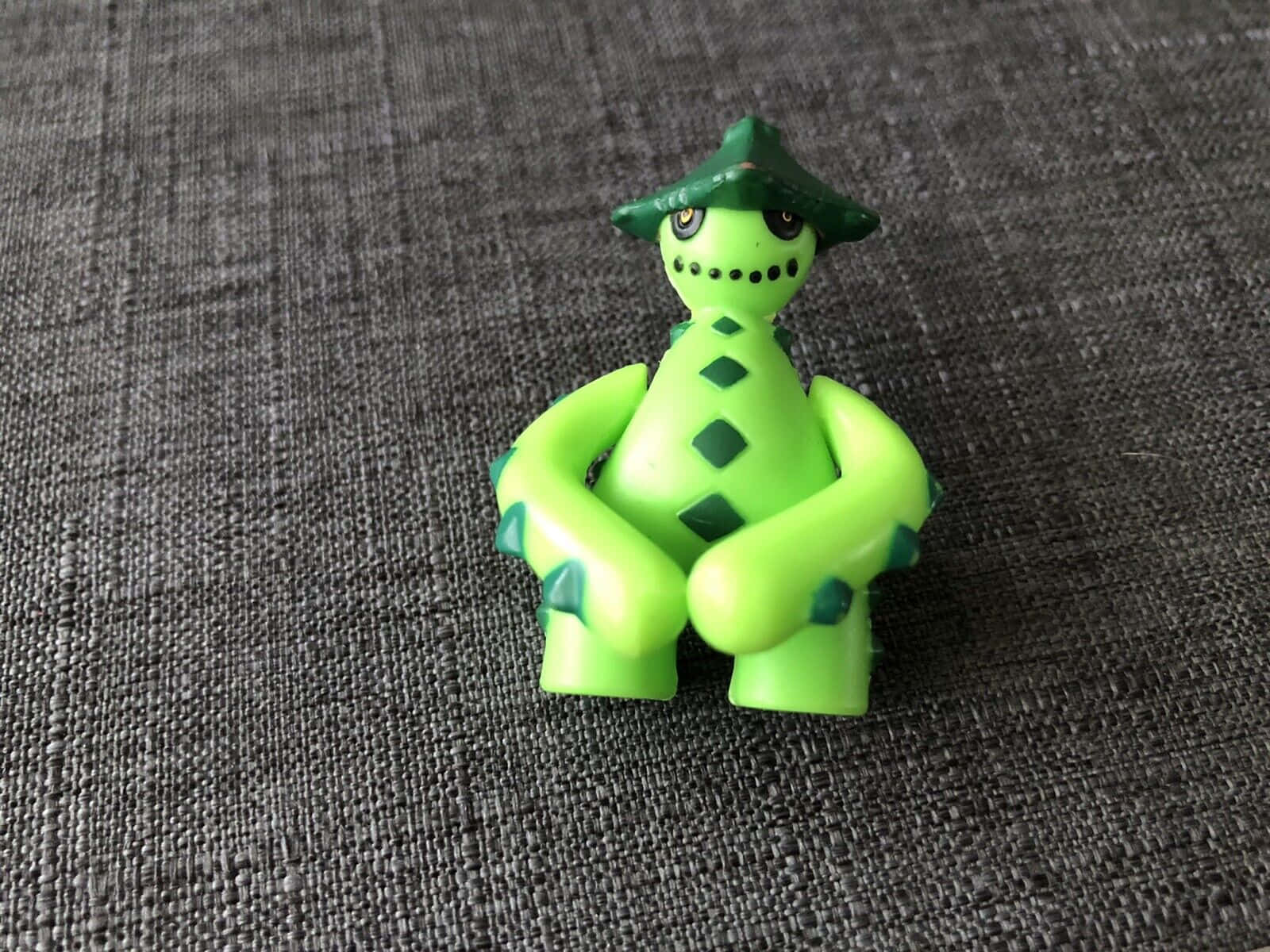 一個戴帽子的綠色塑料玩具人偶盤腿坐在灰色布料上。
