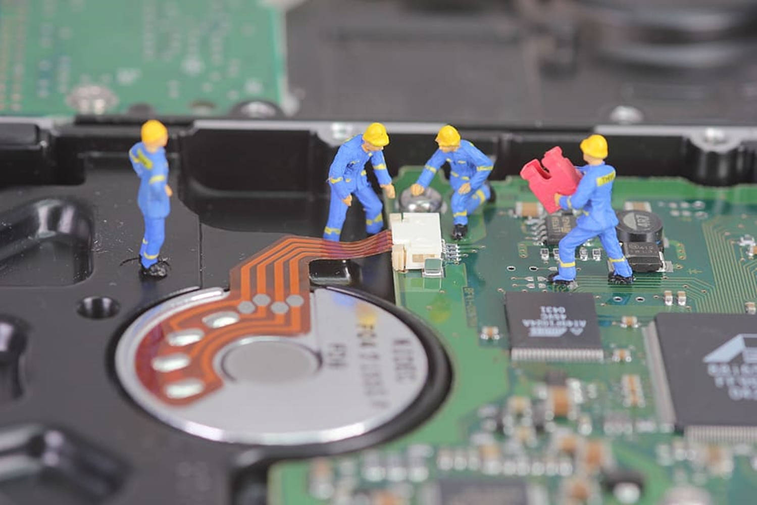 迷你玩具修復電腦硬碟 壁紙 — 身穿藍色制服和黃色頭盔的微型建築工人正在修理硬碟內的綠色電路板。