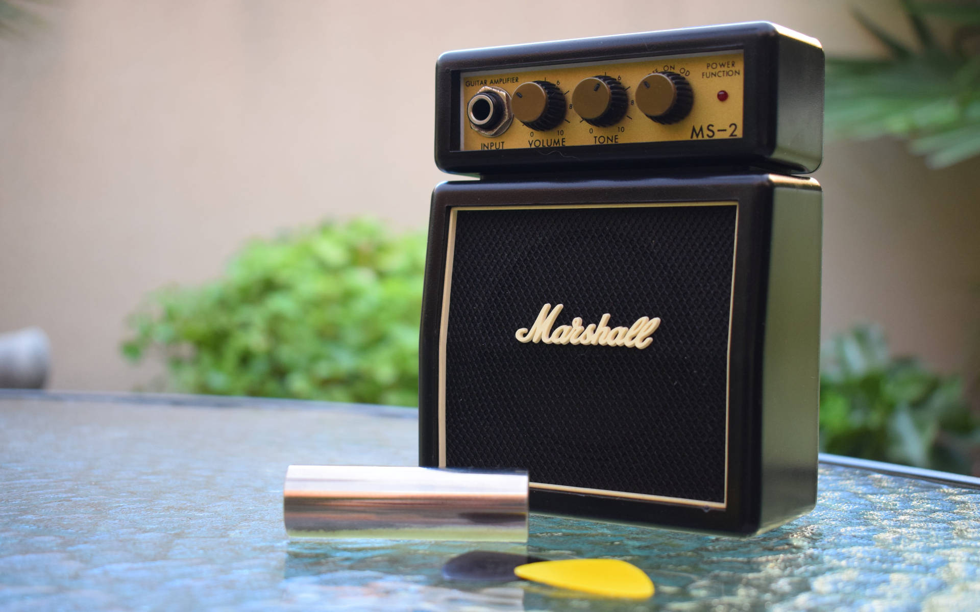 迷你馬歇爾擴大機置於桌面 壁紙 — 一部復古黑色Marshall MS-2結他擴音器放在玻璃桌上，旁邊有黃色撥片和銀色管子。