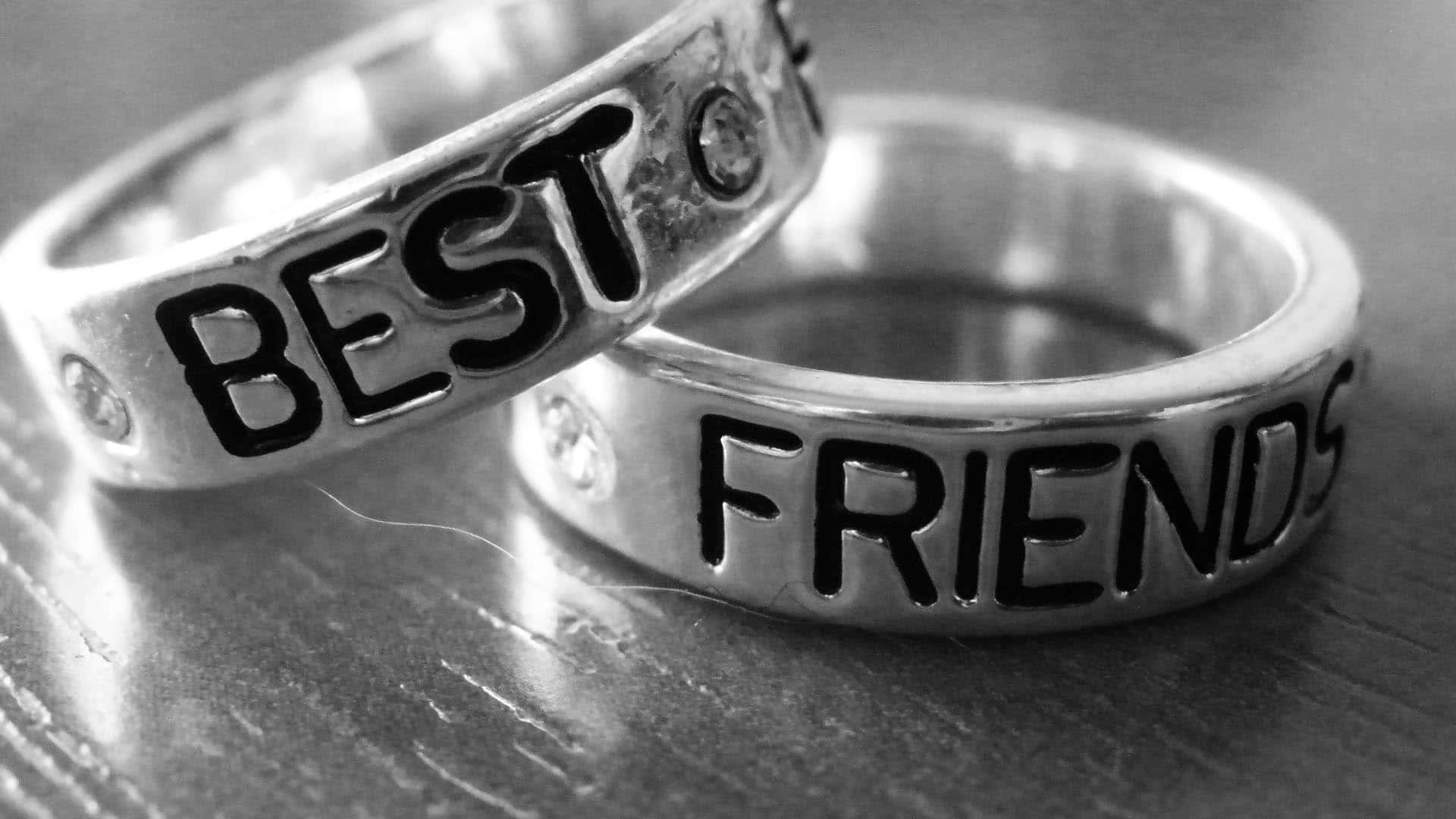 追劇總有回報——與你的「老友記」聚在一起 壁紙 — 兩枚刻有「BEST FRIENDS」字樣的銀戒指，在單色特寫中放置在木質表面上。