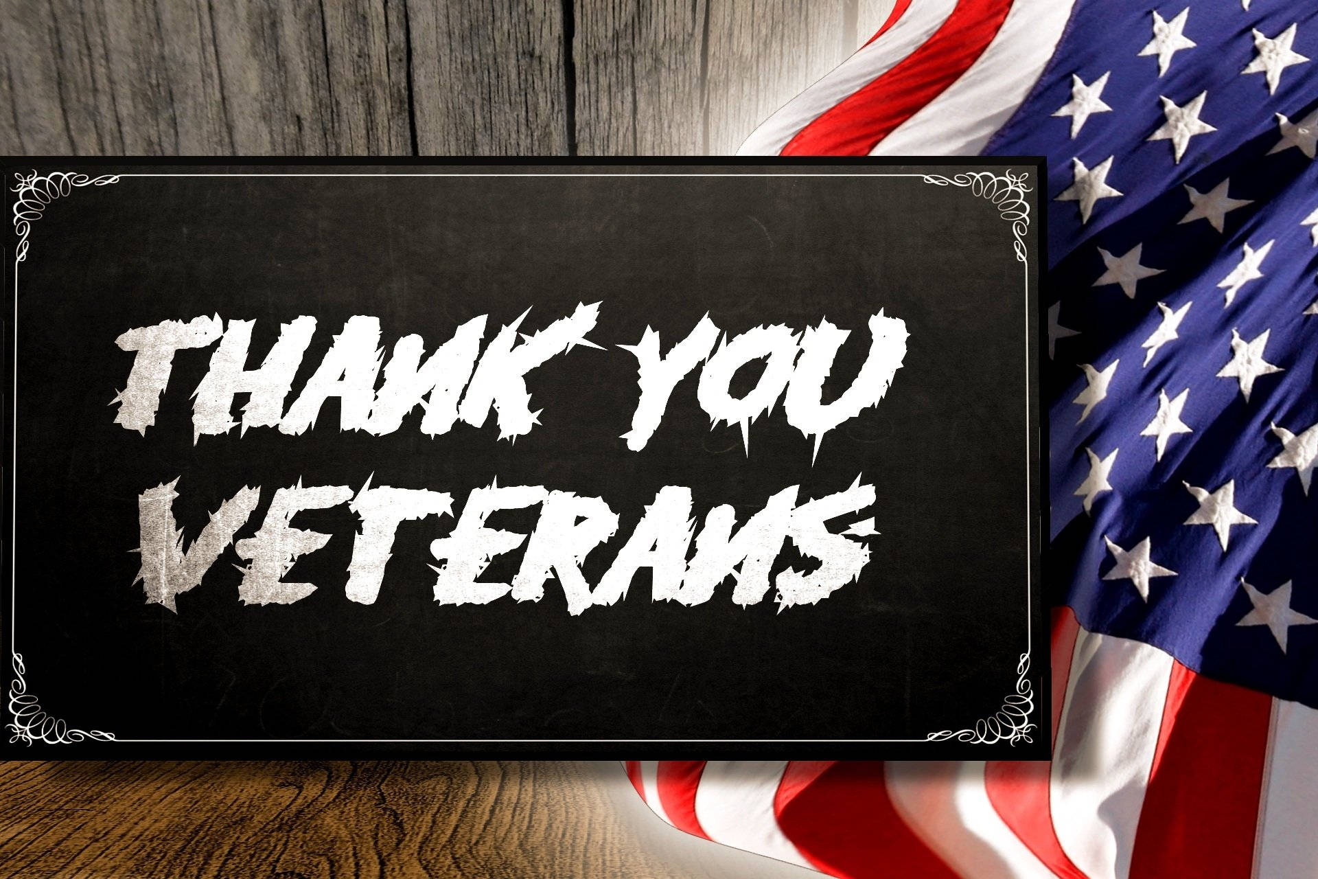 退伍軍人日感謝訊息 壁紙 — 一塊黑板，上面有粗體白色「THANK YOU VETERANS」文字，放在木質表面上，旁邊是垂掛的美國國旗。