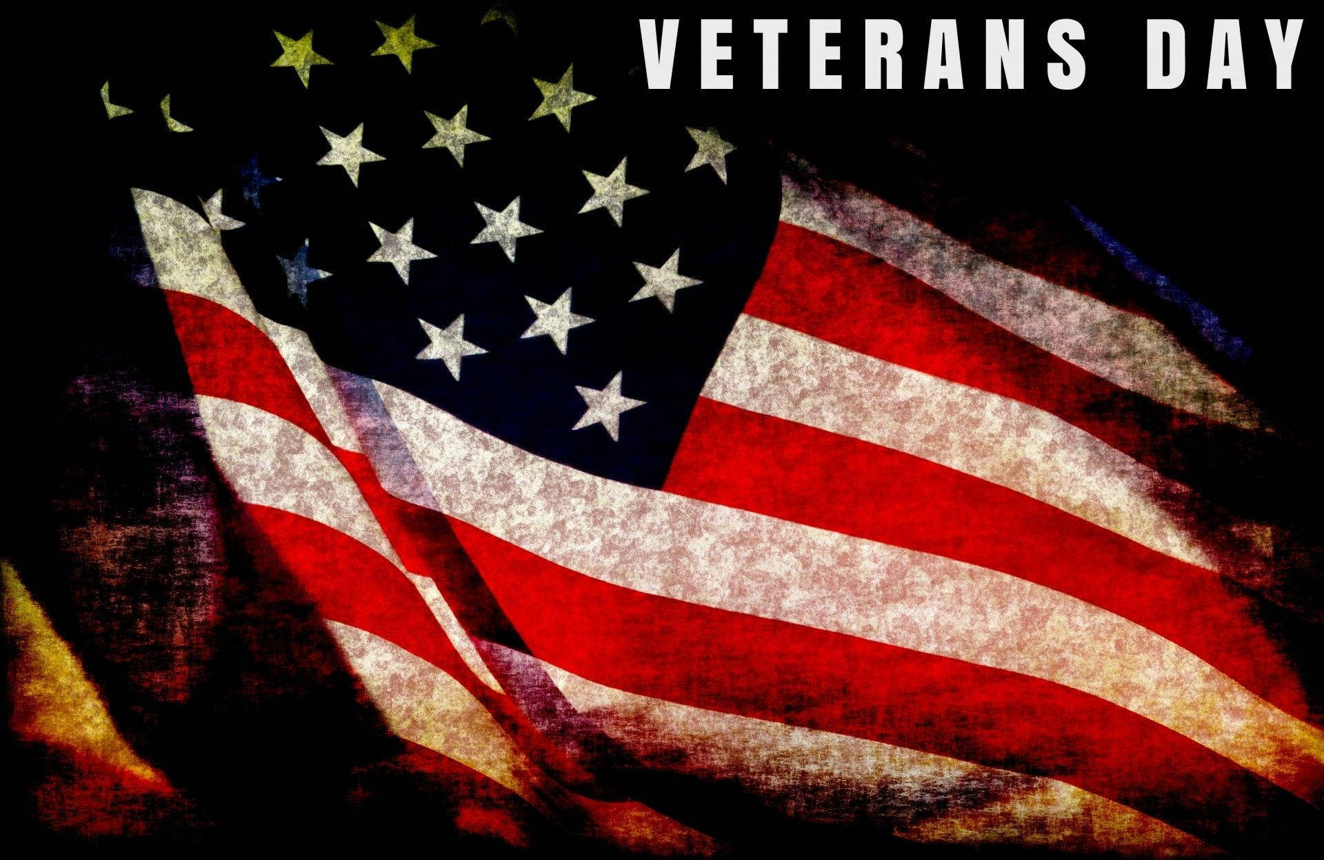 退伍軍人日旗幟與文字 壁紙 — 一面經風化的美國國旗，帶有鮮明的紅、白、藍條紋和星星，在深色紋理背景上飄揚，並帶有「VETERANS DAY」文字。