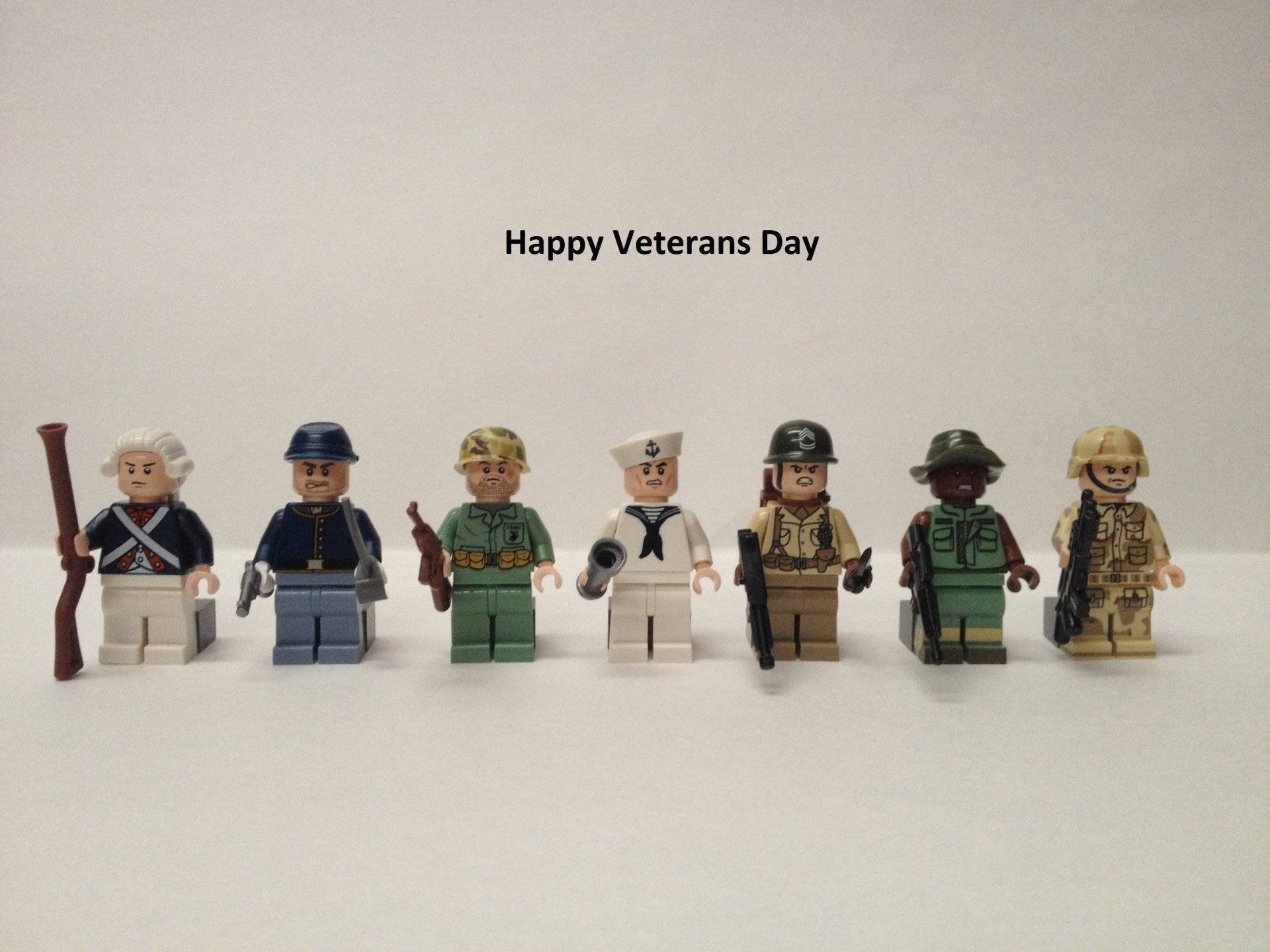 退伍軍人日樂高積木 壁紙 — 七個身穿不同軍服的LEGO士兵人偶排成一排，背景為純白色，上方有「Happy Veterans Day」字樣。