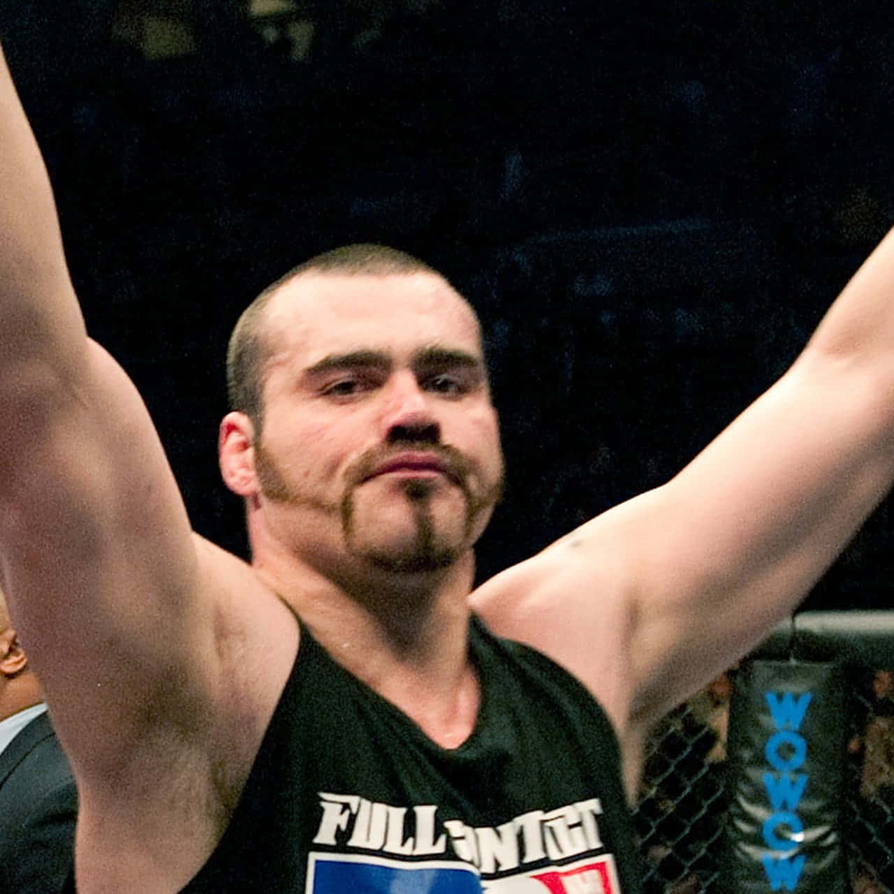 退役綜合格鬥選手 Tim Sylvia 的近照 壁紙 — 一位肌肉發達、留著山羊鬍的鬥士在昏暗的MMA籠中高舉雙臂慶祝勝利。