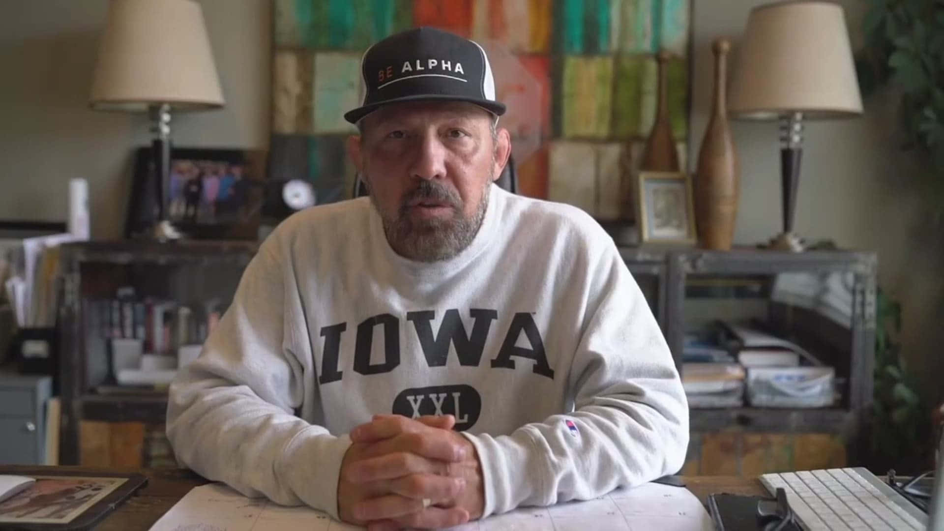 退役綜合格鬥選手帕特·米利蒂奇 壁紙 — 一位留鬍鬚的男子穿著愛荷華州衛衣和be alpha帽，坐在光線溫暖的家庭辦公室中雜亂的書桌前。