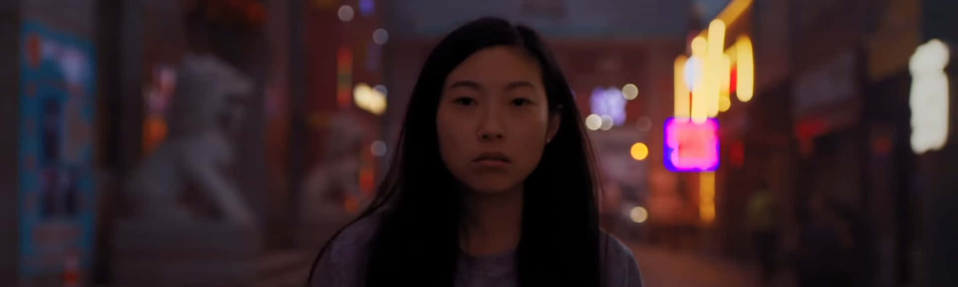 《送別》電影沉思時刻 壁紙 — 一位長黑髮的年輕女性夜晚站在昏暗的城市街道上，周圍是發光的霓虹燈招牌和模糊的商店櫥窗。
